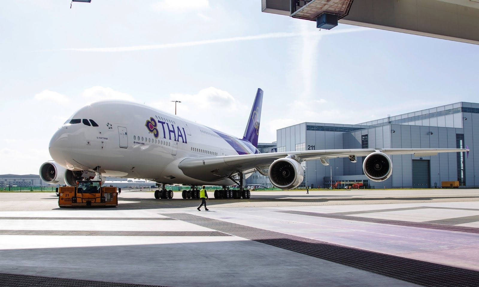 飛機 桌布 壁紙 — 一架泰國航空的空中巴士A380正被一輛地面車輛拖過陽光普照的機場停機坪，附近有一名工作人員。
