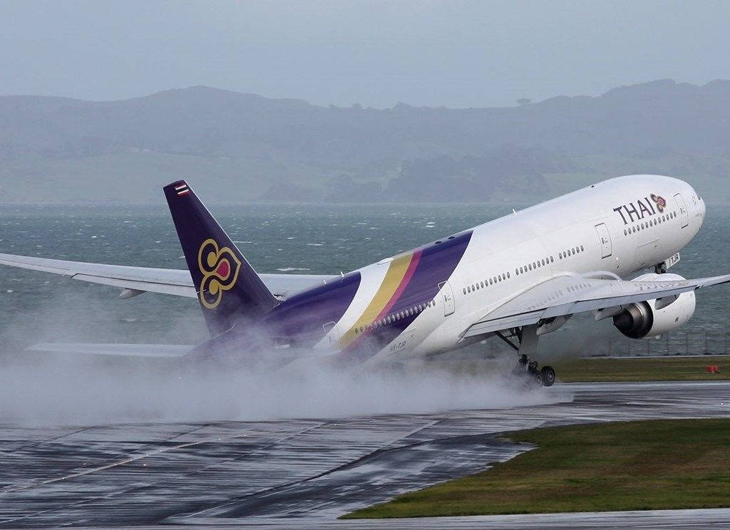 飛機 桌布 壁紙 — 一架泰國航空飛機在潮濕的跑道上降落，濺起水花，雨水落在遠處的山丘和水面上。