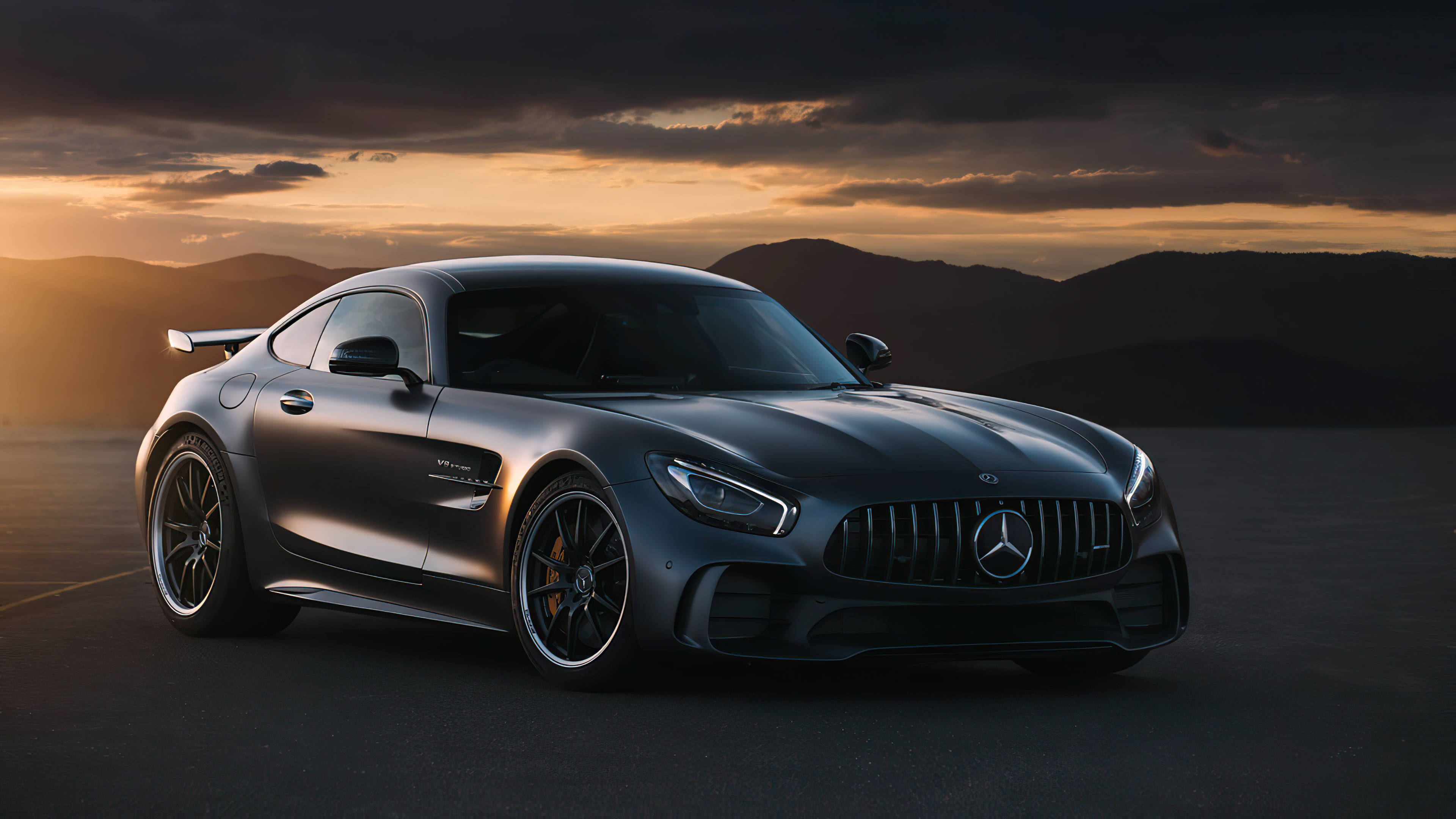 Mercedes Benz Amg 桌布 壁紙 — 一架流線型啞光灰色Mercedes-AMG GT停泊在日落時分的開闊道路上，背景有戲劇性的雲層和遠山。