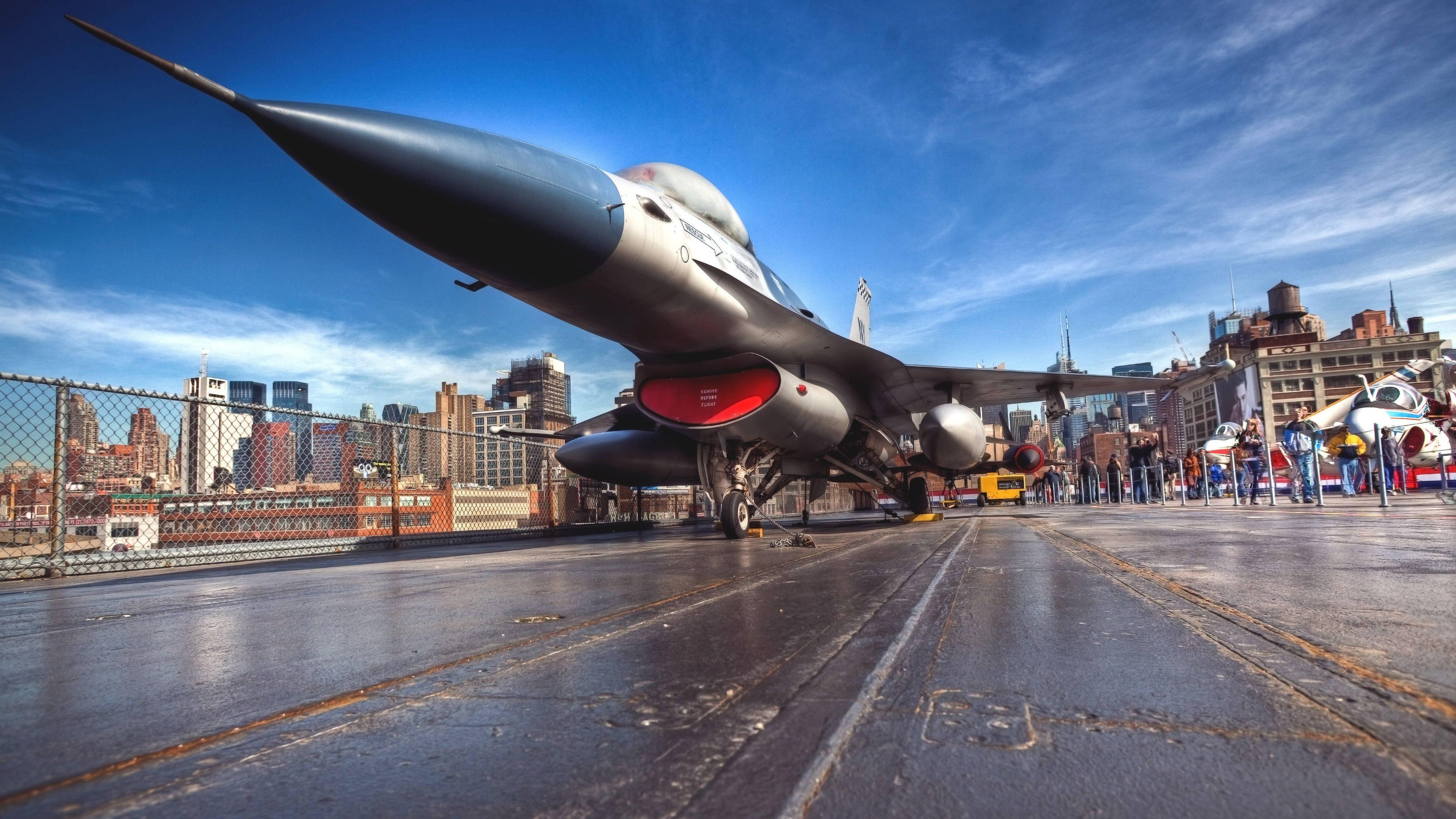 桌布 壁紙 — 一架流線型戰鬥機主導著濕潤的航空母艦甲板，背景是晴朗藍天下充滿活力的城市天際線。