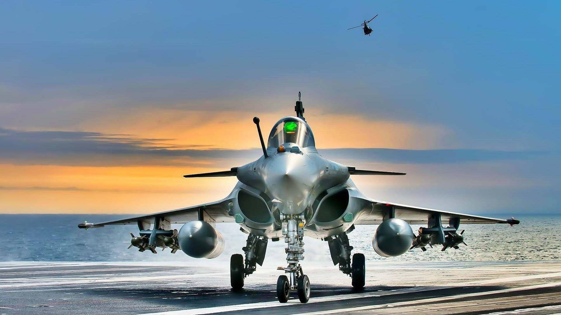 桌布 壁紙 — 一架流線型戰鬥機在日落時分在航空母艦甲板上滑行，一架直升機在上空飛行。
