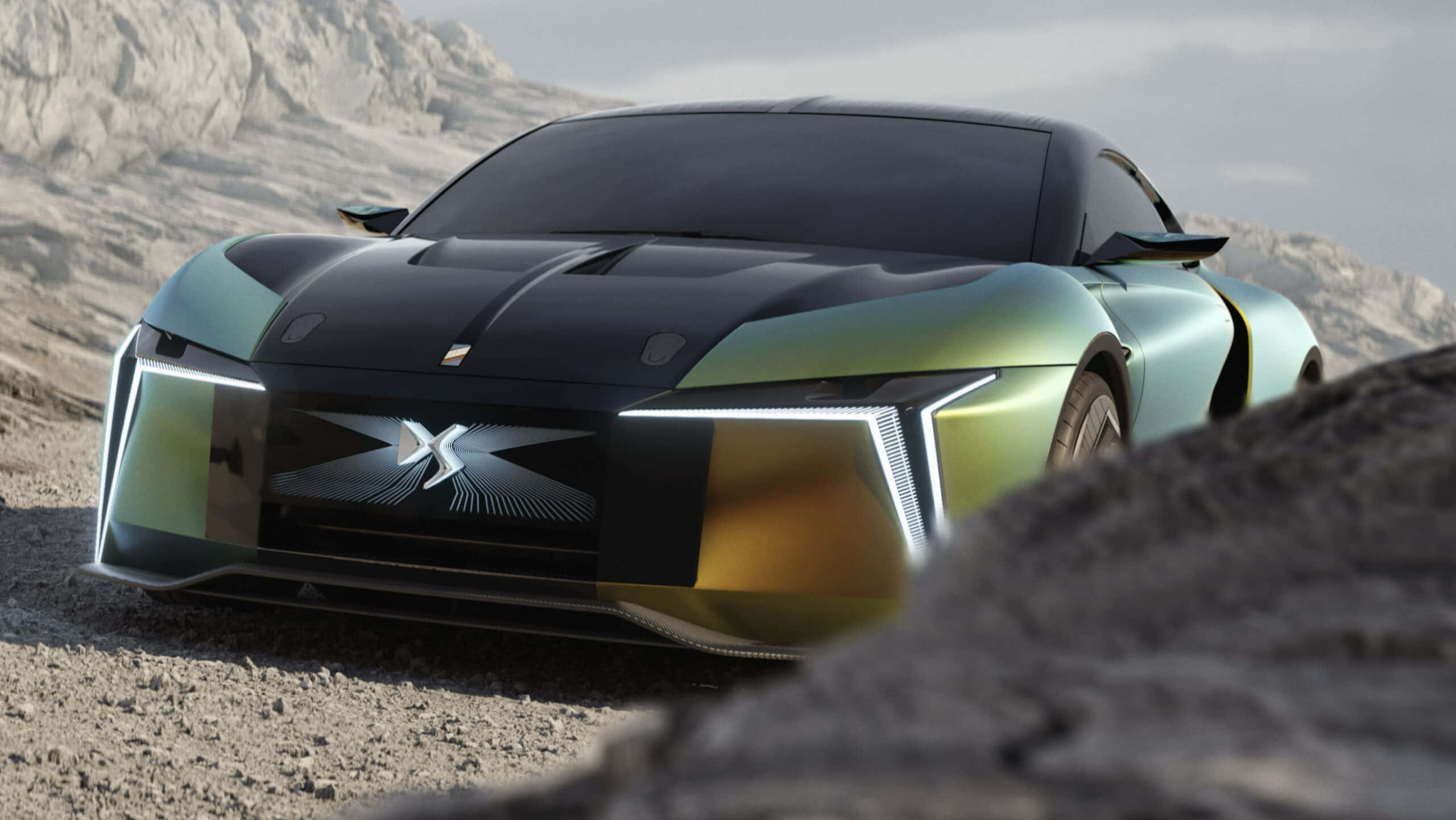 Ds Automobiles 桌布 壁紙 — 一架流線型未來感跑車，帶有彩虹色綠色和黑色塗裝，停放在岩石沙漠地形上，背景有山脈。
