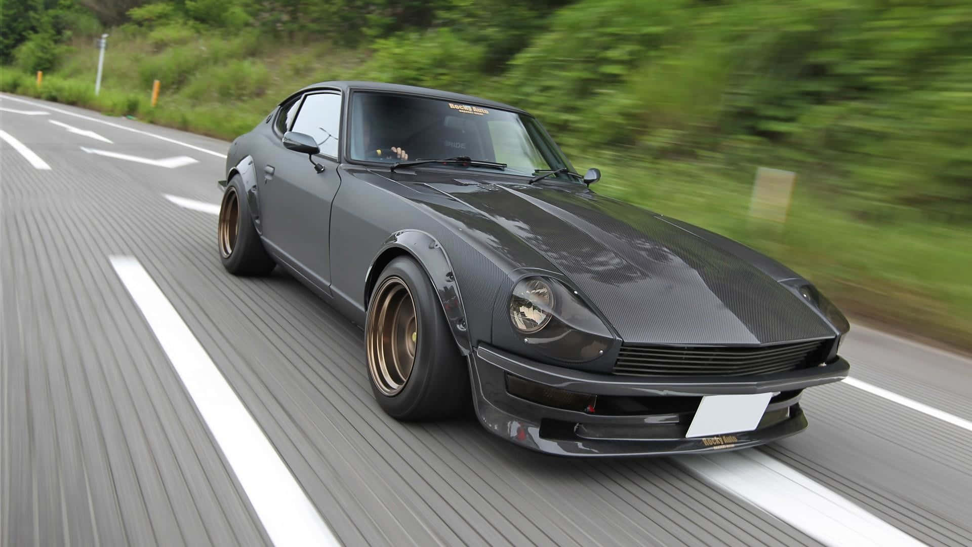 Datsun 桌布 壁紙 — 一架流線型灰色Datsun 240Z在蜿蜒的道路上疾馳，茂密的綠樹在運動中模糊掠過。
