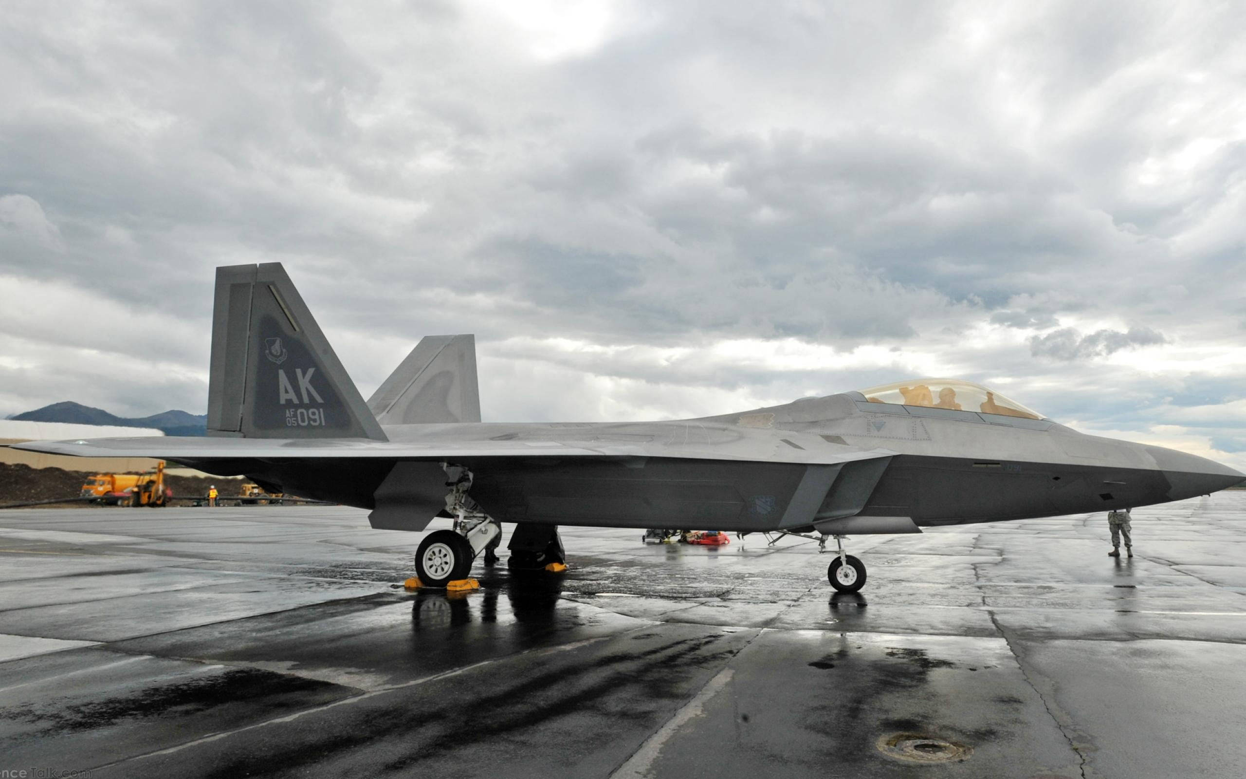 transportation 桌布 壁紙 — 一架流線型灰色F-22猛禽戰鬥機停在潮濕的停機坪上，天空多雲，遠山可見。