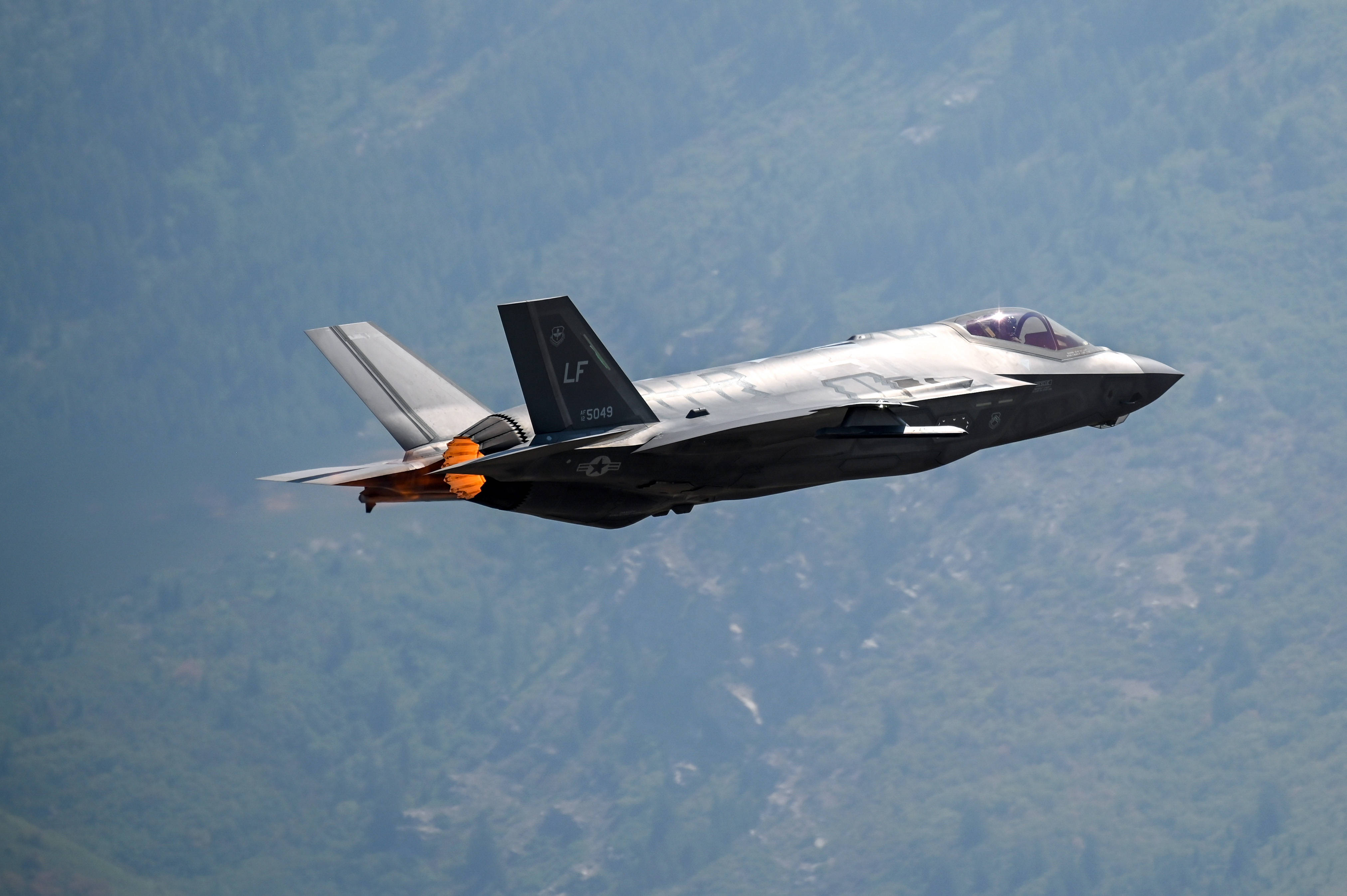 Plane 桌布 壁紙 — 一架流線型灰色F-35戰鬥機低飛過森林覆蓋的山脈，拖著橙色後燃器火焰。