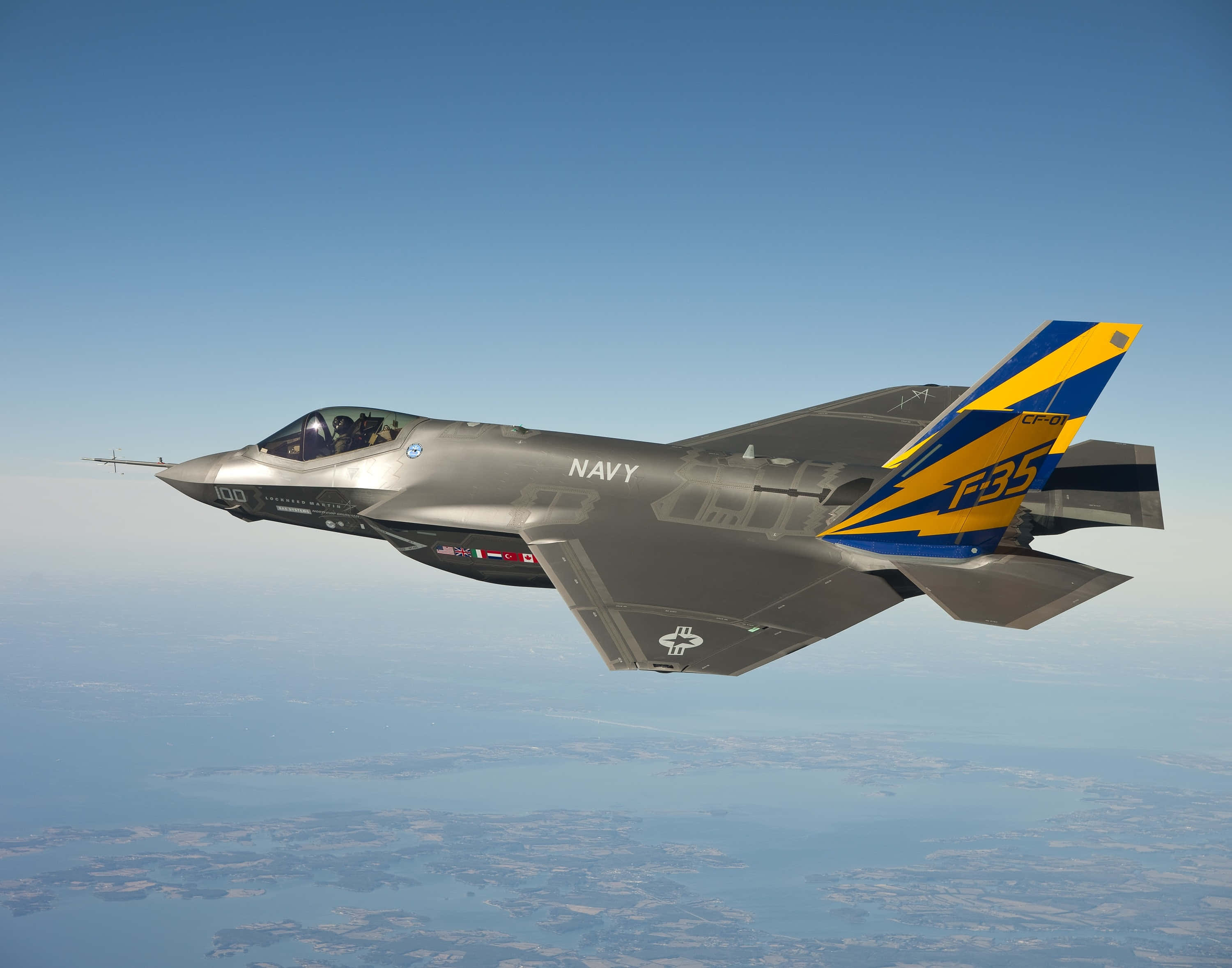 Jet 桌布 壁紙 — 一架流線型灰色F-35戰鬥機帶有藍黃色尾翼標誌，在晴朗藍天下翱翔於海岸景觀之上。