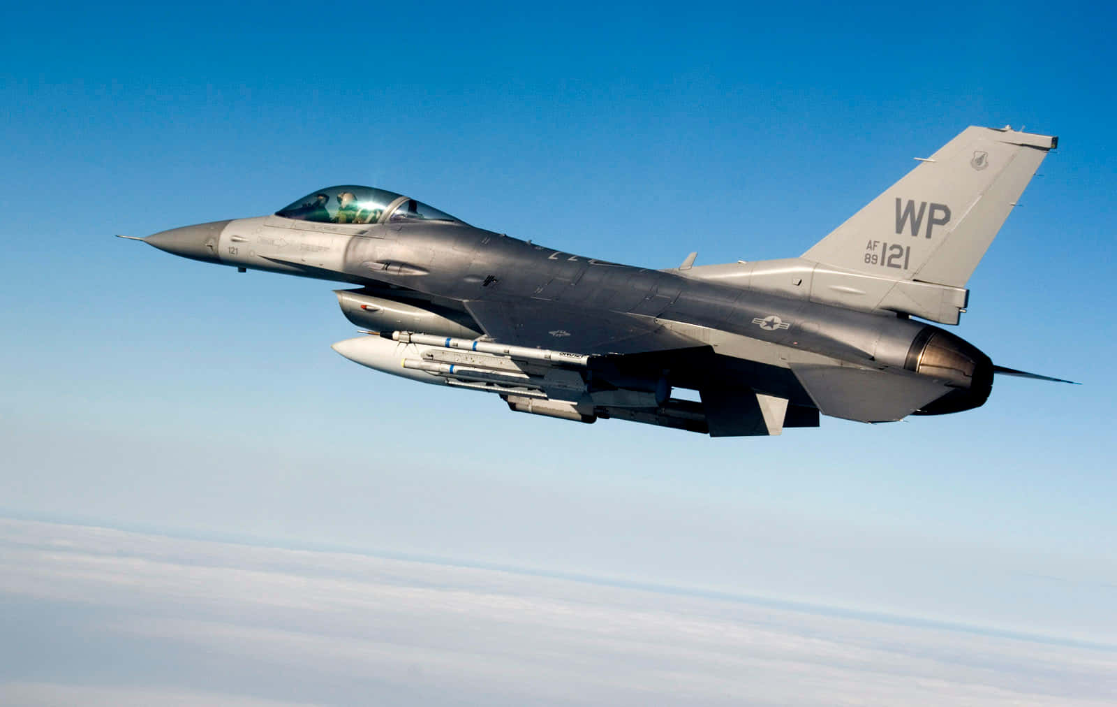 Air Force 桌布 壁紙 — 一架流線型灰色戰鬥機在晴朗藍天下翱翔於蓬鬆白雲之上，可見飛行員及武器裝備。