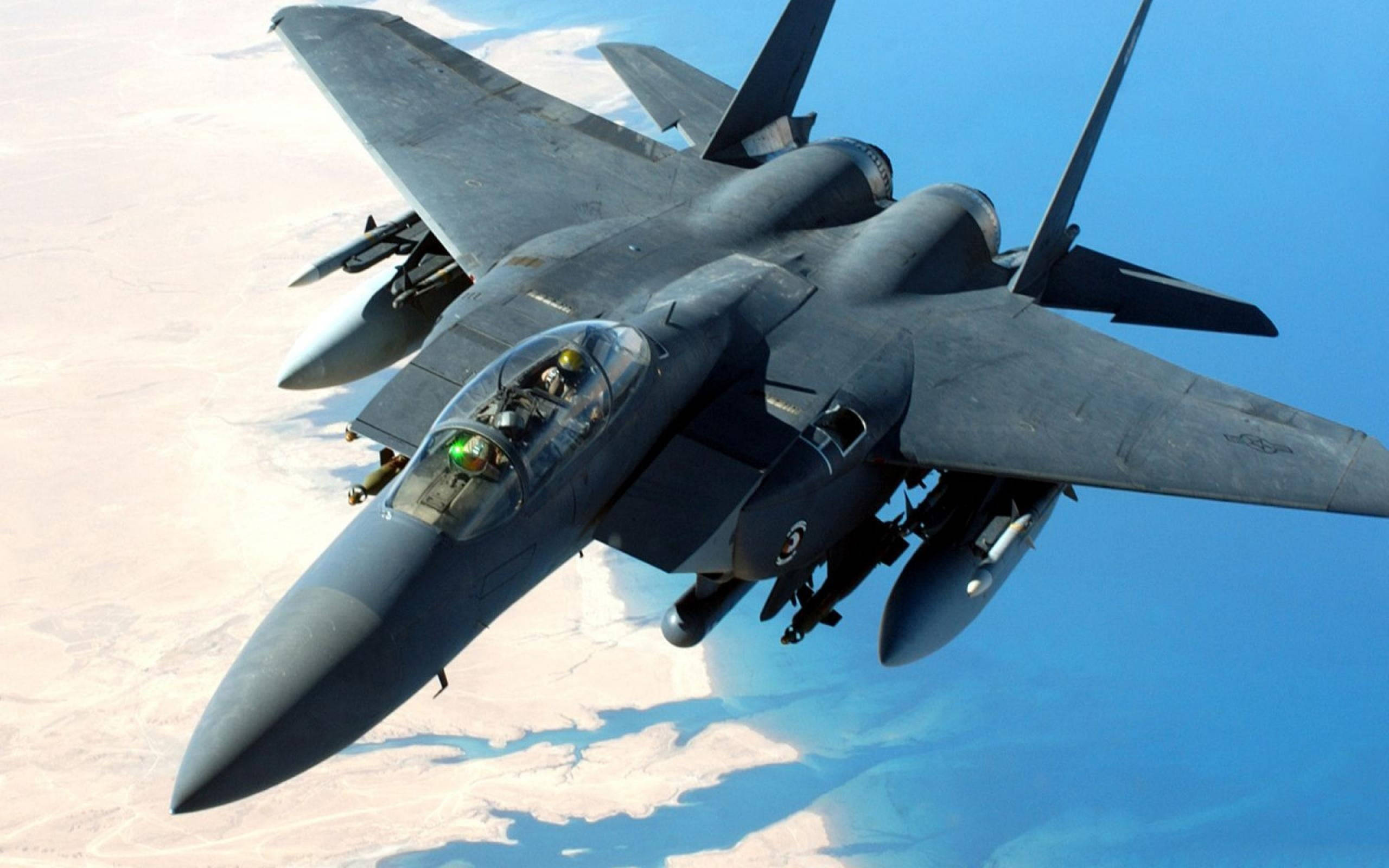 軍事動圖 桌布 壁紙 — 一架流線型灰色戰鬥機在晴朗藍天下翱翔於沙漠景觀之上，駕駛艙內的飛行員清晰可見。