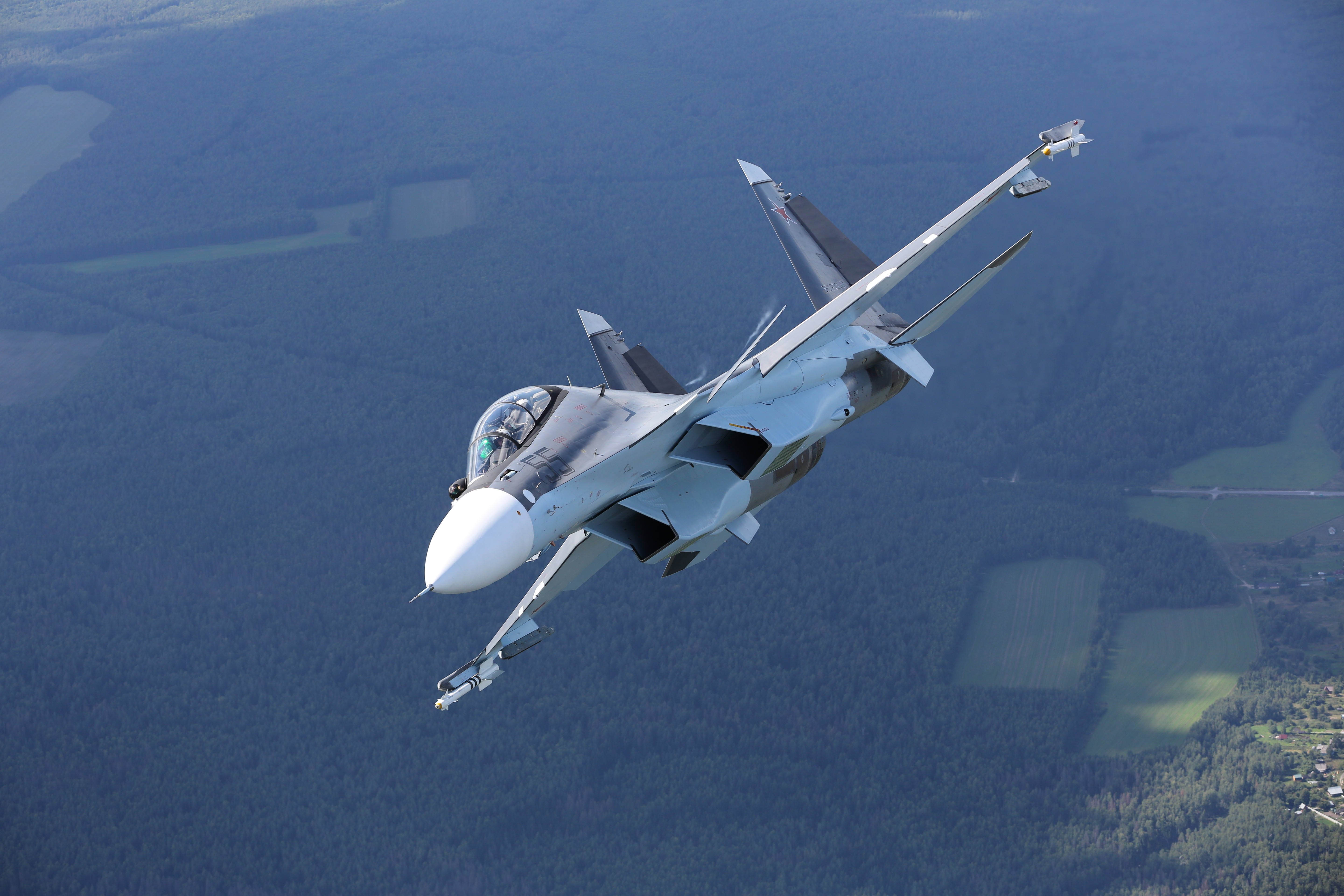 Air Force 桌布 壁紙 — 一架流線型灰色戰鬥機在森林景觀和下方綠色田野上空動態翱翔。