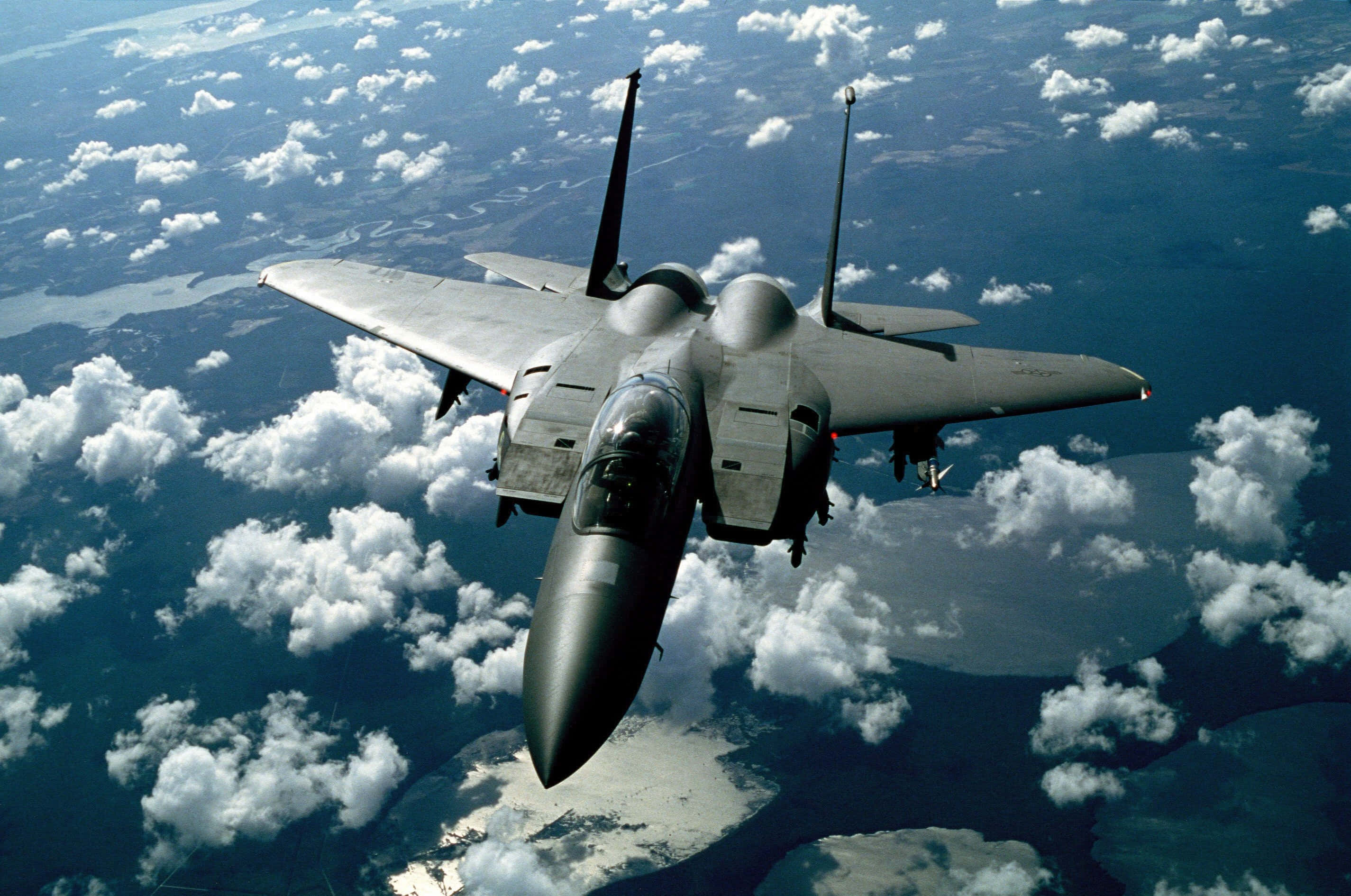 Fighter Jet 桌布 壁紙 — 一架流線型灰色戰鬥機在蓬鬆的白雲上方翱翔，下方是藍色海洋和陸地。