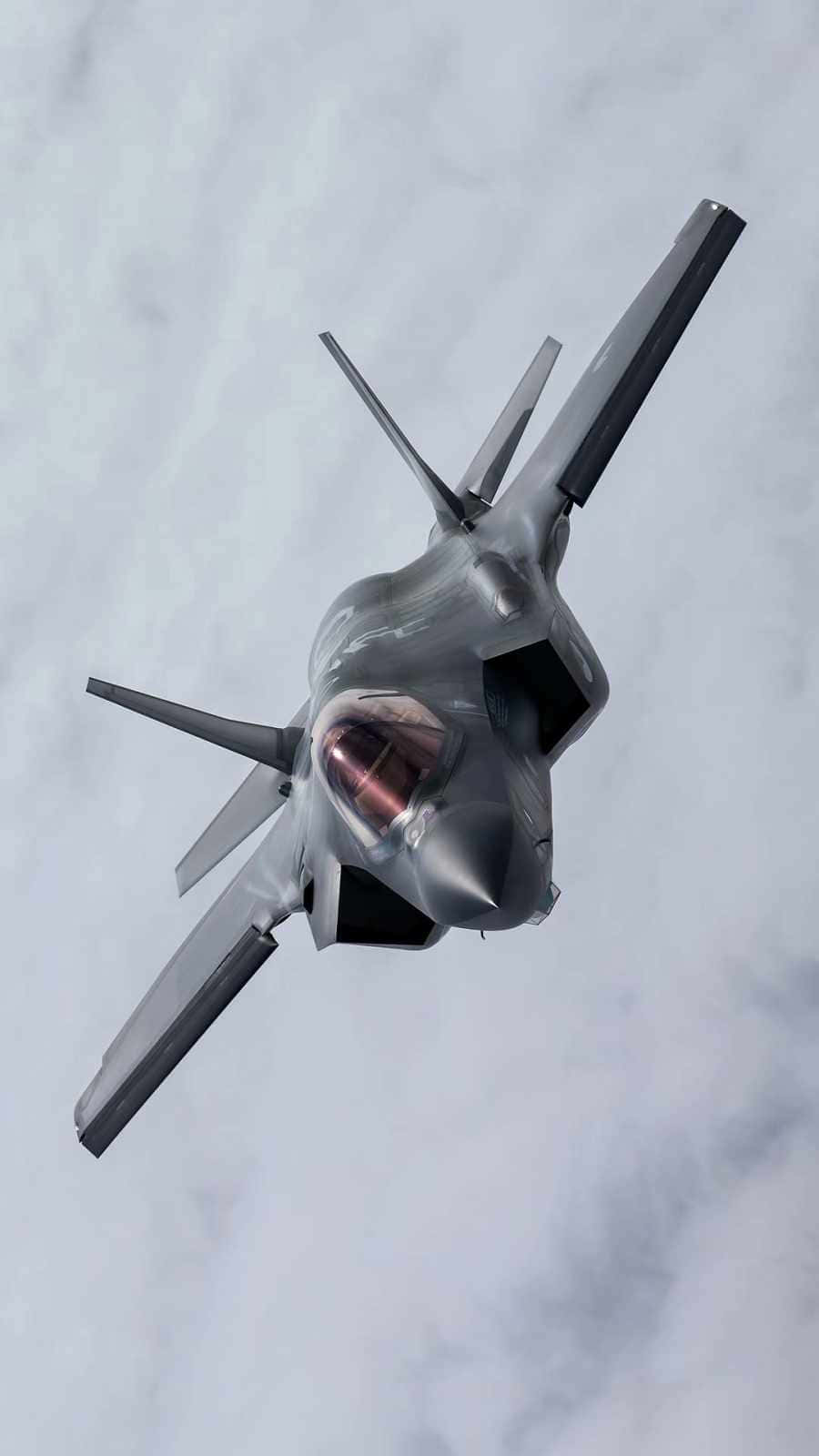 Android Jumbo Jets 桌布 壁紙 — 一架流線型灰色隱形戰鬥機在多雲的天空中翱翔，駕駛艙發出溫暖的光芒。
