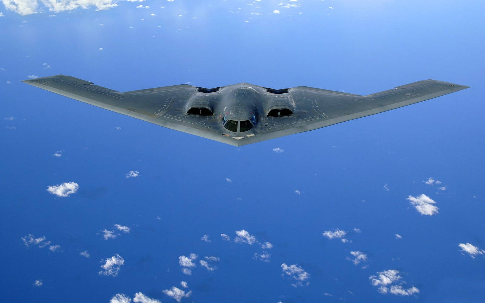 Air Force 桌布 壁紙 — 一架流線型灰色隱形轟炸機在廣闊的藍天中翱翔，點綴著散落的白色雲朵。