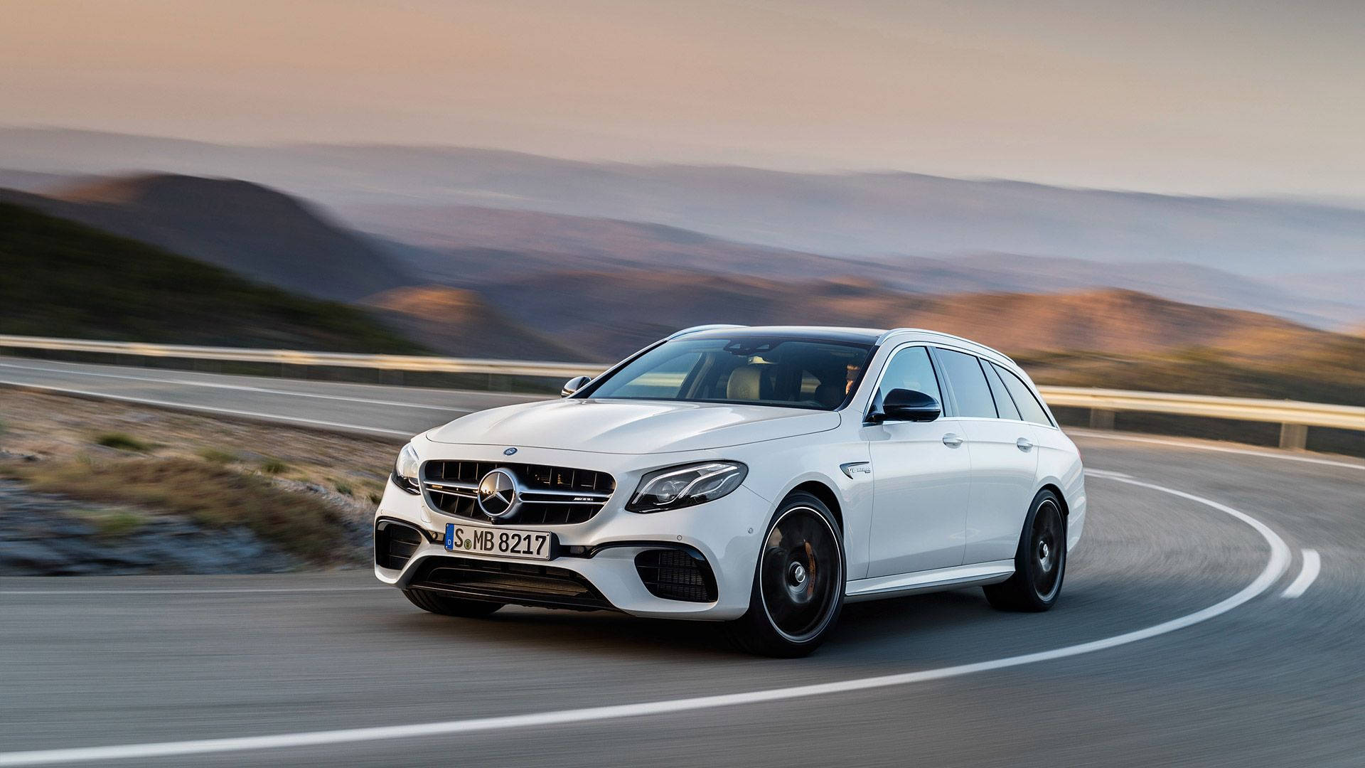 Mercedes 桌布 壁紙 — 一架流線型白色 Mercedes-AMG E63 S Estate 在日落時分沿著蜿蜒的山路疾馳，背景是模糊的山丘。