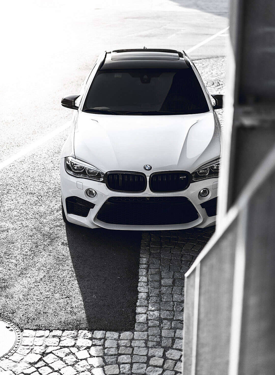 黑白高畫質 桌布 壁紙 — 一架流線型白色BMW SUV從上方拍攝，停泊在鵝卵石街道上，強烈的陰影和城市建築構成了畫面的框架。
