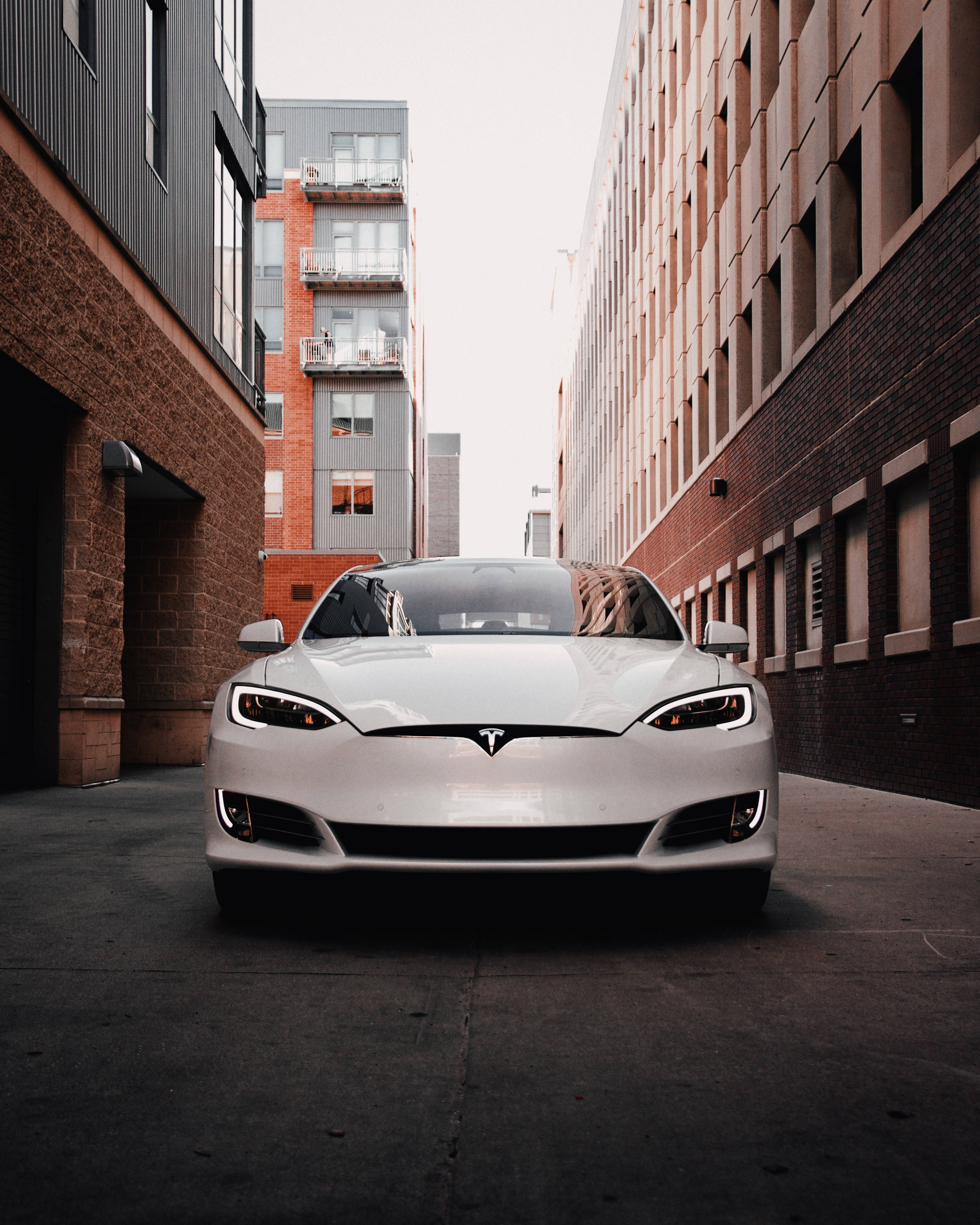 特斯拉 桌布 壁紙 — 一架流線型白色Tesla Model S在狹窄的城市小巷中向前，兩旁是磚砌和現代建築，天空多雲。
