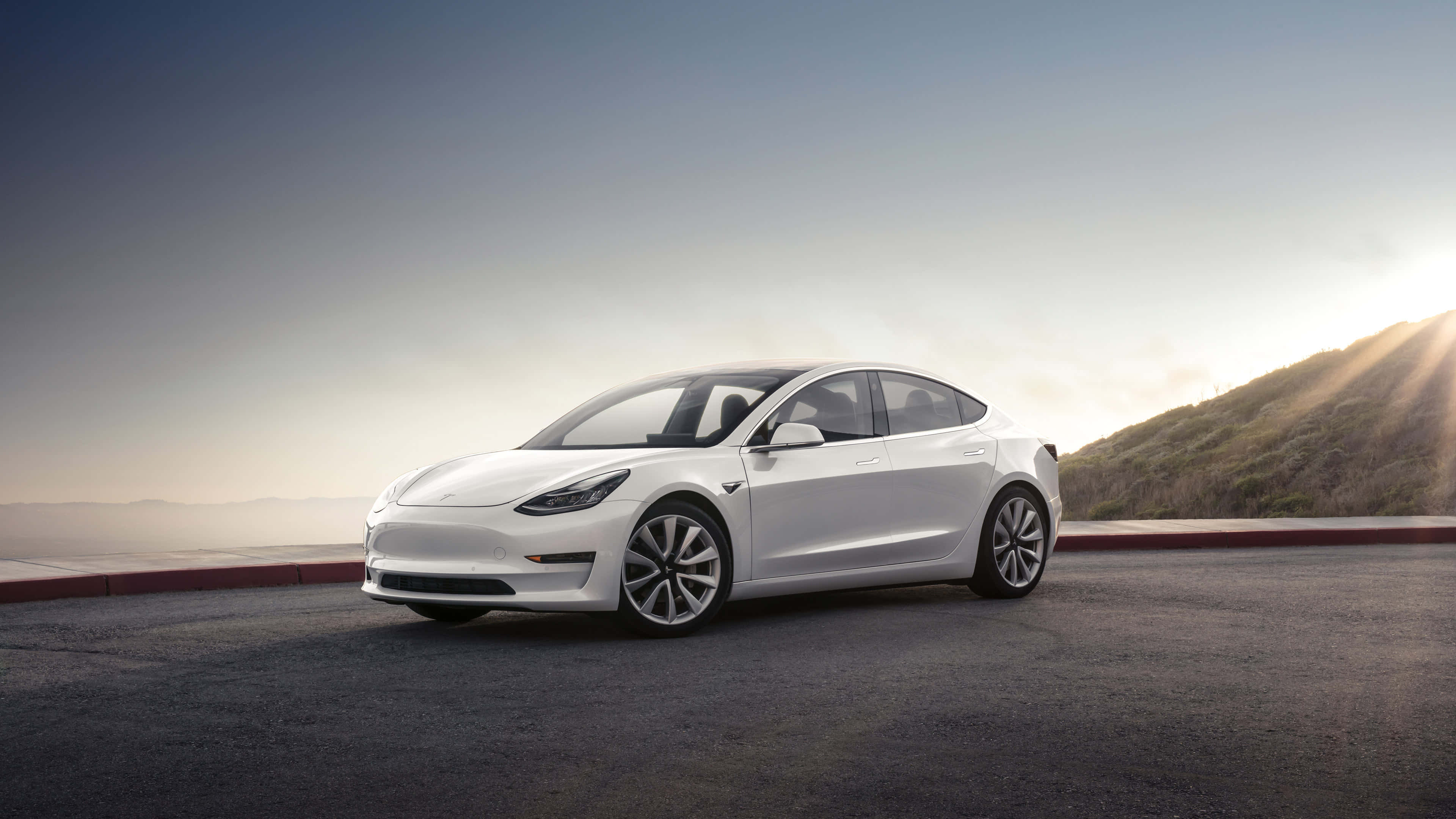 Tesla Model 3 桌布 壁紙 — 一架流線型白色Tesla Model 3停泊在路邊，俯瞰著黃金時段陽光照耀的山坡。
