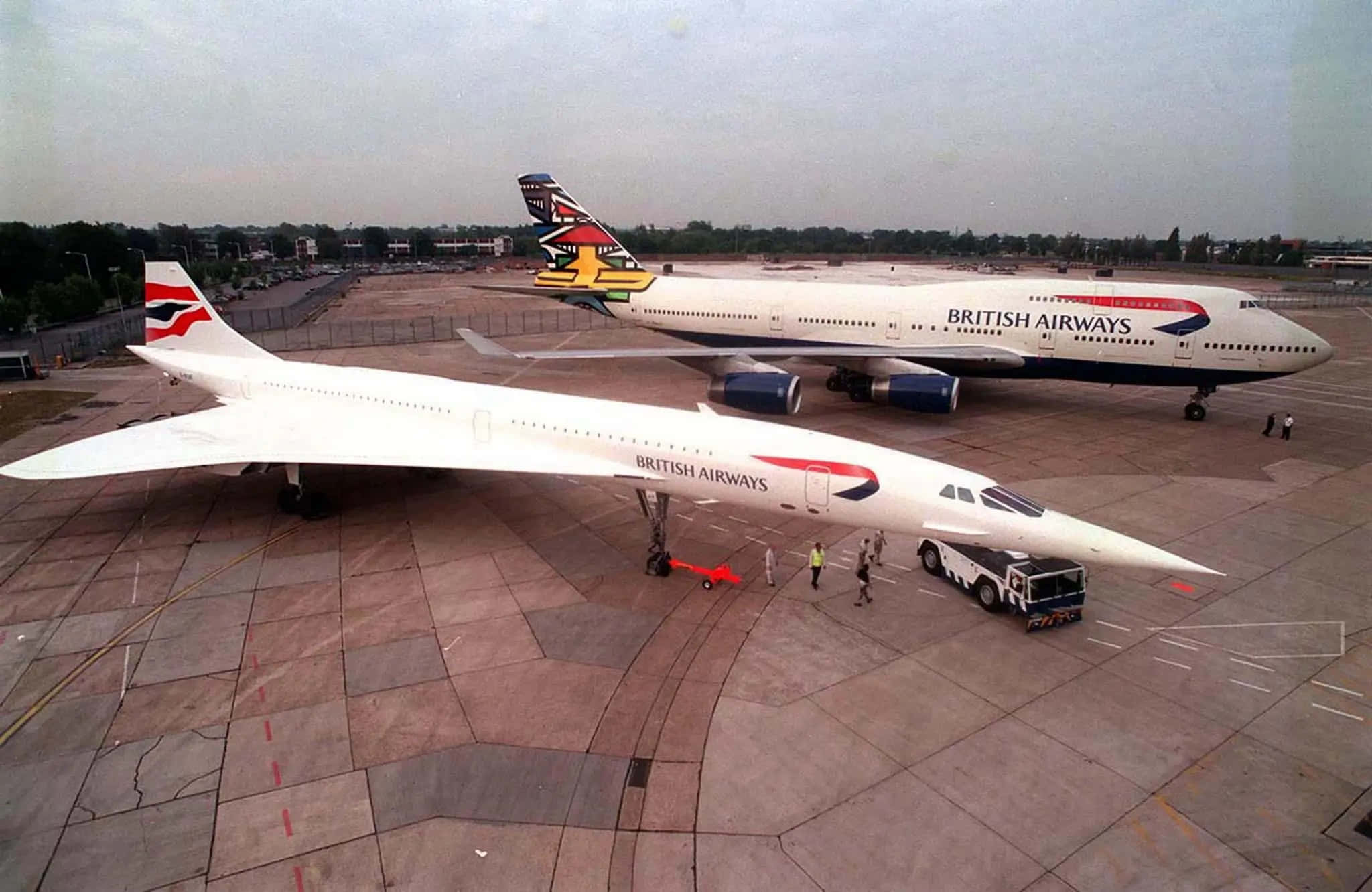 Jumbo Jets 桌布 壁紙 — 一架流線型白色協和客機與一架英國航空波音747並排停在停機坪上，附近有地勤人員。