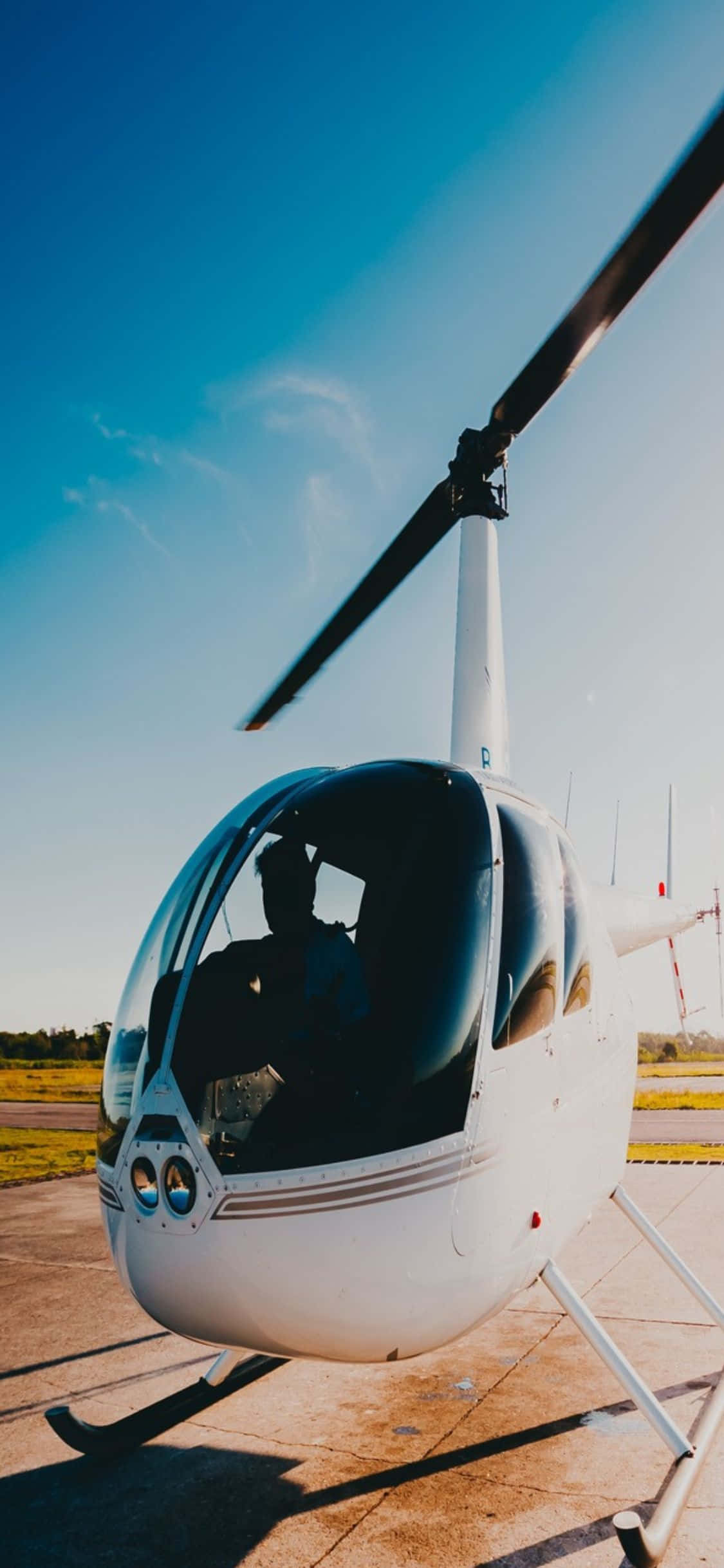 iPhone Xs Helicopters 桌布 壁紙 — 一架流線型白色直升機停在停機坪上，天空清澈湛藍，柔和陽光投下長長的影子。