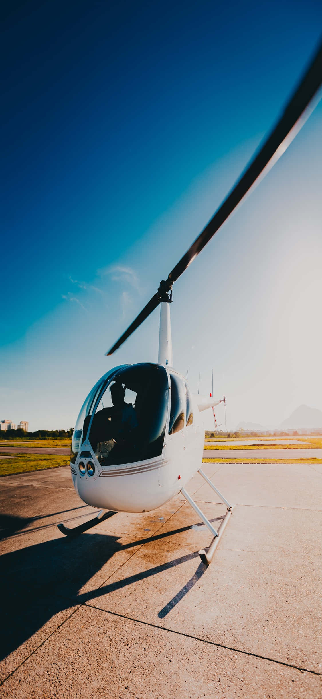 iPhone Xs Helicopters 桌布 壁紙 — 一架流線型白色直升機停在陽光普照的停機坪上，藍天白雲，長長的影子在混凝土上延伸。