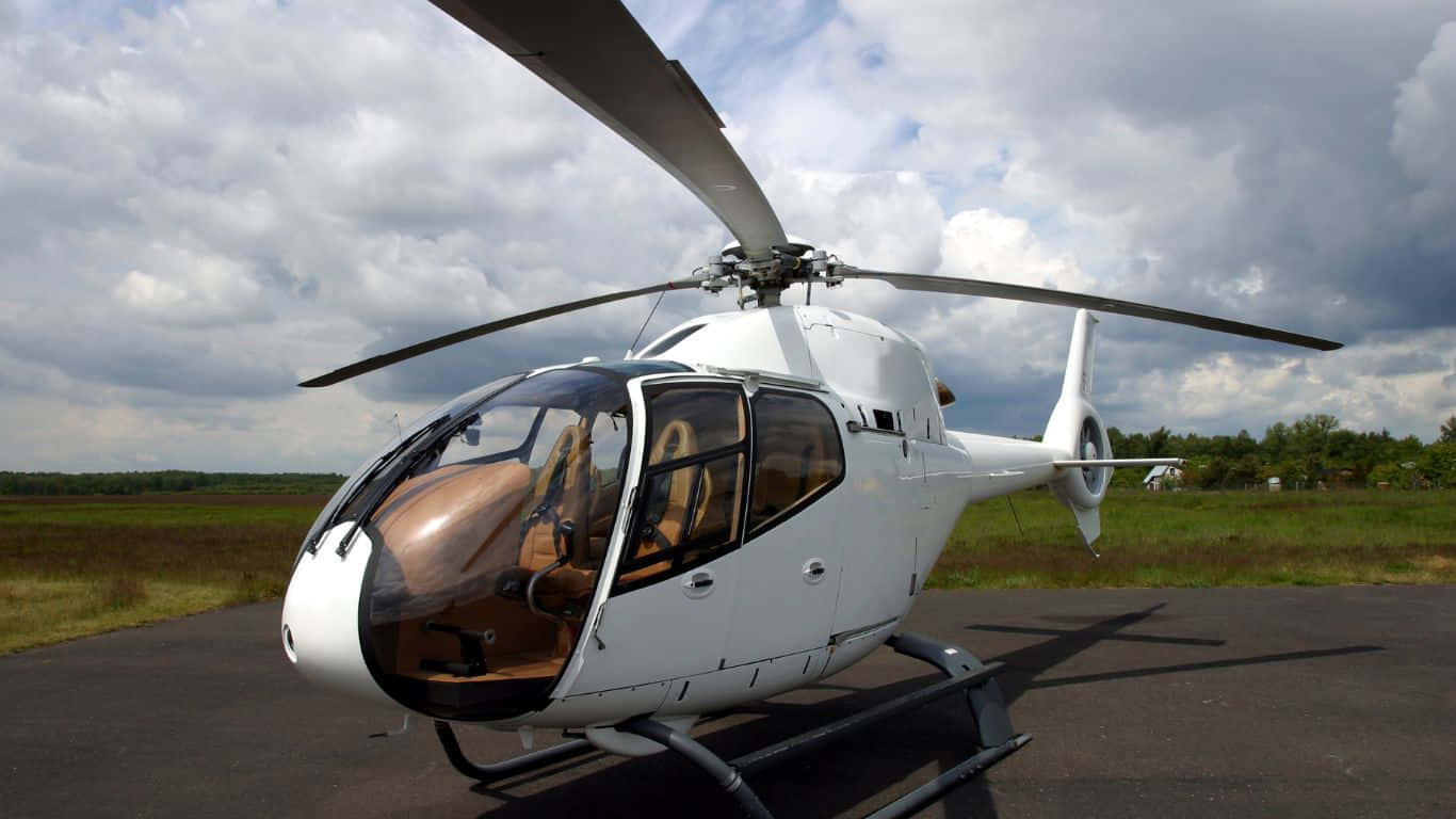 1366x768 Helicopters 桌布 壁紙 — 一架流線型白色直升機停泊在鋪砌的停機坪上，天空多雲，背景是綠色的田野。