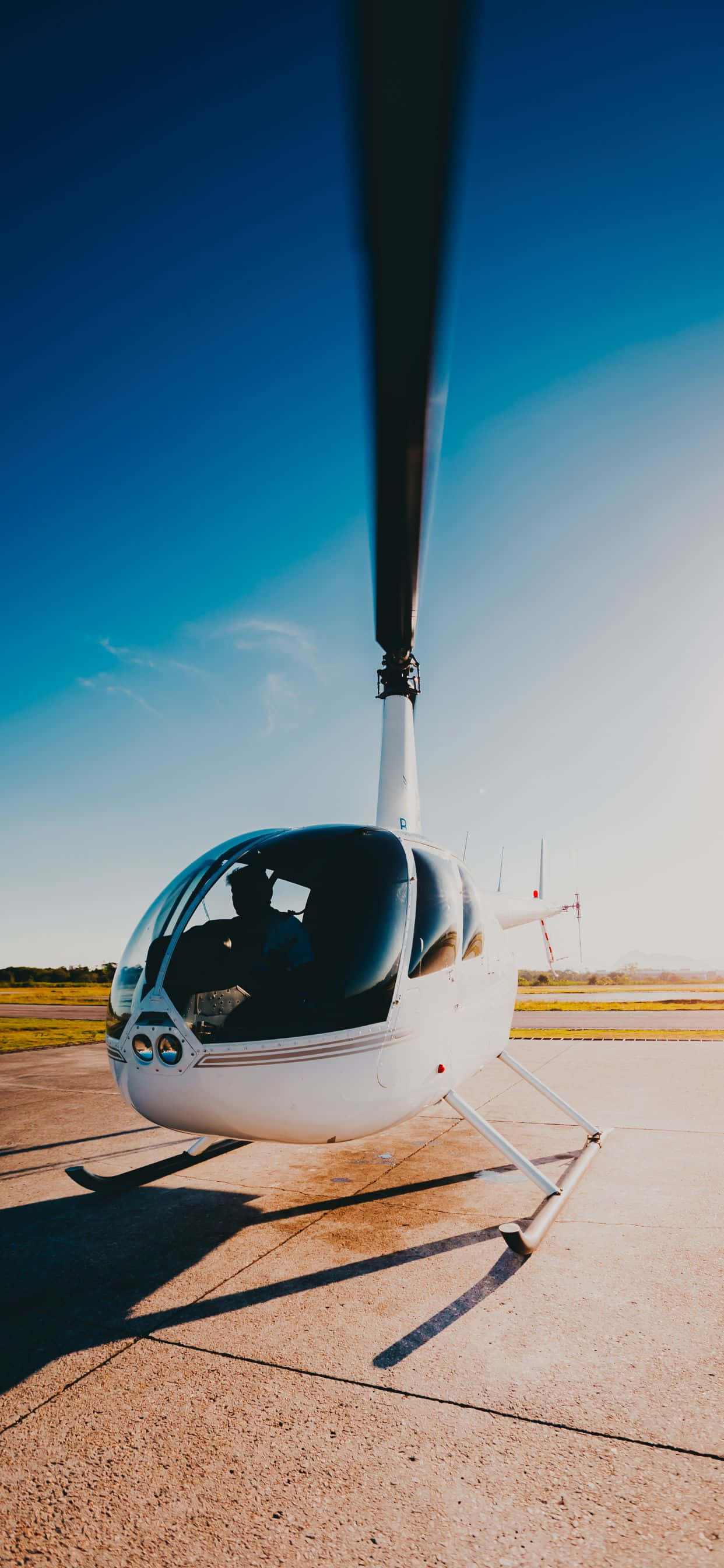iPhone Xs Max Helicopters 桌布 壁紙 — 一架流線型白色直升機停泊在陽光普照的停機坪上，背景是廣闊清澈的藍天，旋翼葉片投下長長的影子。