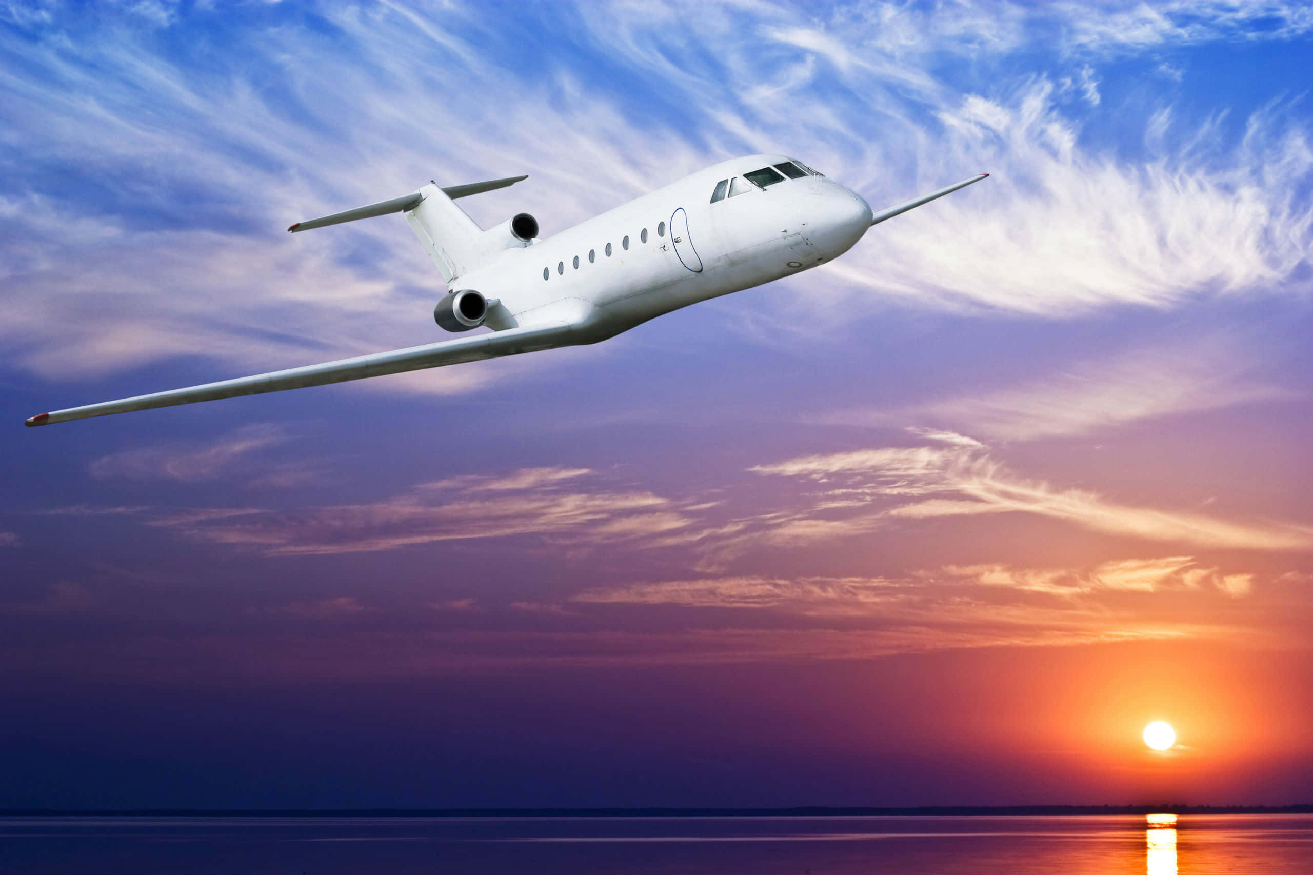 HD Plane 桌布 壁紙 — 一架流線型白色私人飛機在平靜的海平線上空，穿過充滿活力的日落天空和縷縷雲彩。