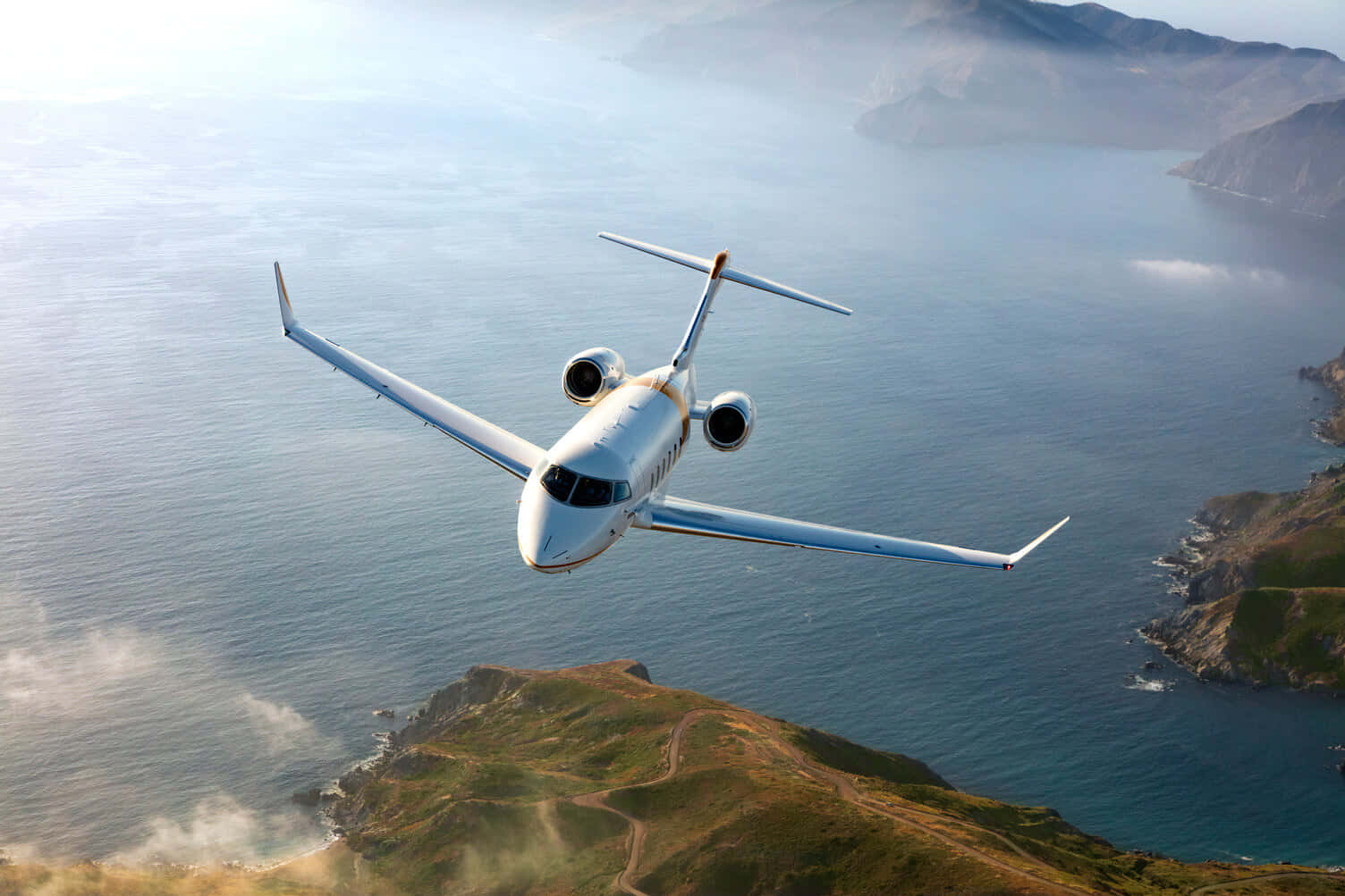 飛機 桌布 壁紙 — 一架流線型白色私人飛機在霧氣瀰漫的海岸景觀上空翱翔，下方是深藍色的海洋和崎嶇的綠色懸崖。