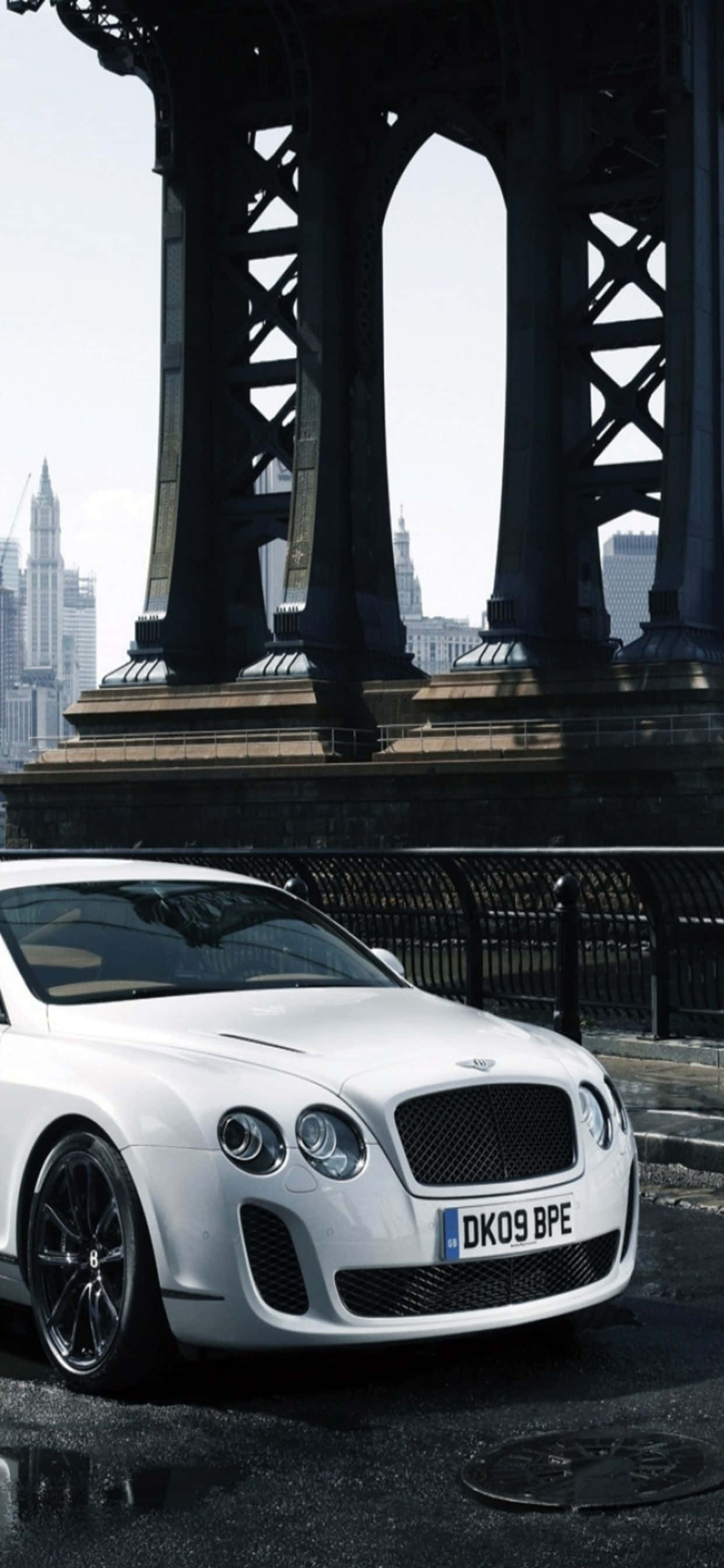 iPhone Xs Max Bentley 桌布 壁紙 — 一架流線型白色賓利Continental GT停泊在巨型鋼橋的陰影下，背景是城市天際線。