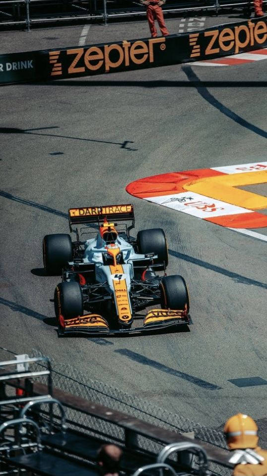 Racing 桌布 壁紙 — 一架流線型的橙色和銀色一級方程式賽車在陽光明媚的賽道上奔馳，賽道有紅色、白色和黃色的路肩，背景中可見穿著橙色工作服的維修站人員。
