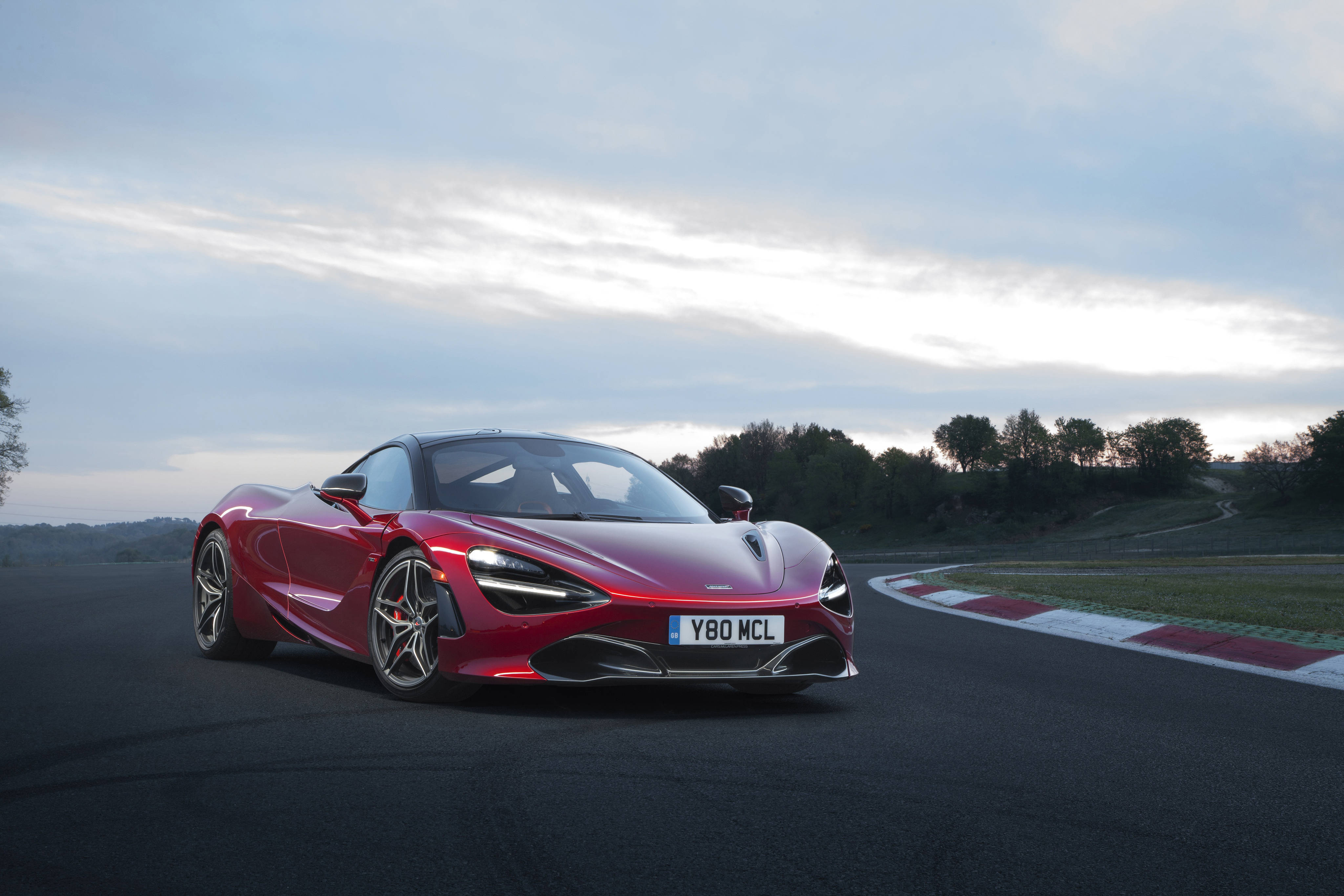 mclaren 720s 4K 桌布 壁紙 — 一架流線型的紅色McLaren 720S跑車在黃昏時分穿梭於蜿蜒的賽道上，背景有樹木和多雲的天空。
