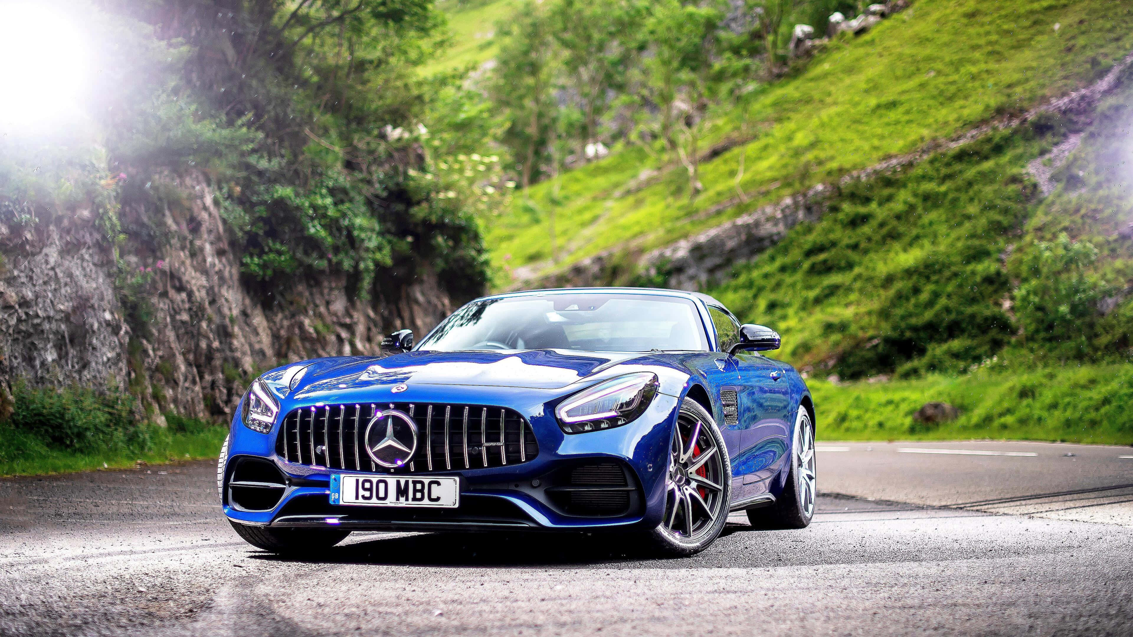 Mercedes Amg 桌布 壁紙 — 一架流線型的藍色 Mercedes-AMG GT 跑車在蜿蜒的山路上閃閃發光，周圍是翠綠的山丘和岩石峭壁。