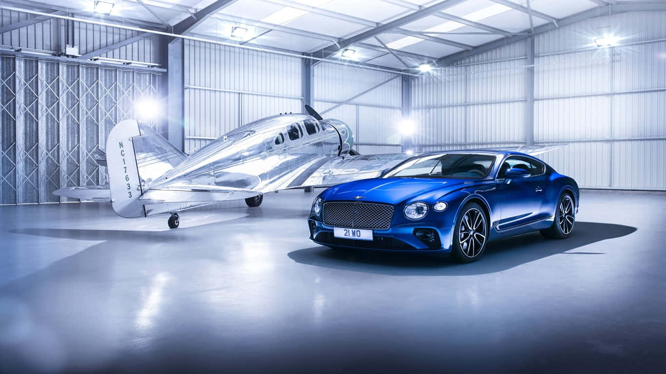 1366x768 Bentley 桌布 壁紙 — 一架流線型的藍色賓利 Continental GT 停在一架銀色復古飛機旁邊，位於明亮的工業機庫內。