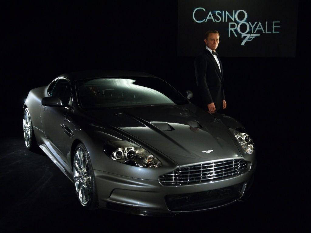 Daniel Craig 桌布 壁紙 — 一架流線型的銀色Aston Martin DBS在攝影棚燈光下閃閃發光，丹尼爾·克雷格身穿禮服站在旁邊，背景為黑色，帶有Casino Royale 75標誌。