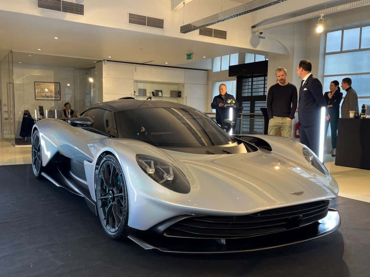 奧斯頓馬丁 桌布 壁紙 — 一架流線型的銀色Aston Martin超跑在明亮燈光下閃閃發光，一群男士在現代豪華展示間中欣賞它。