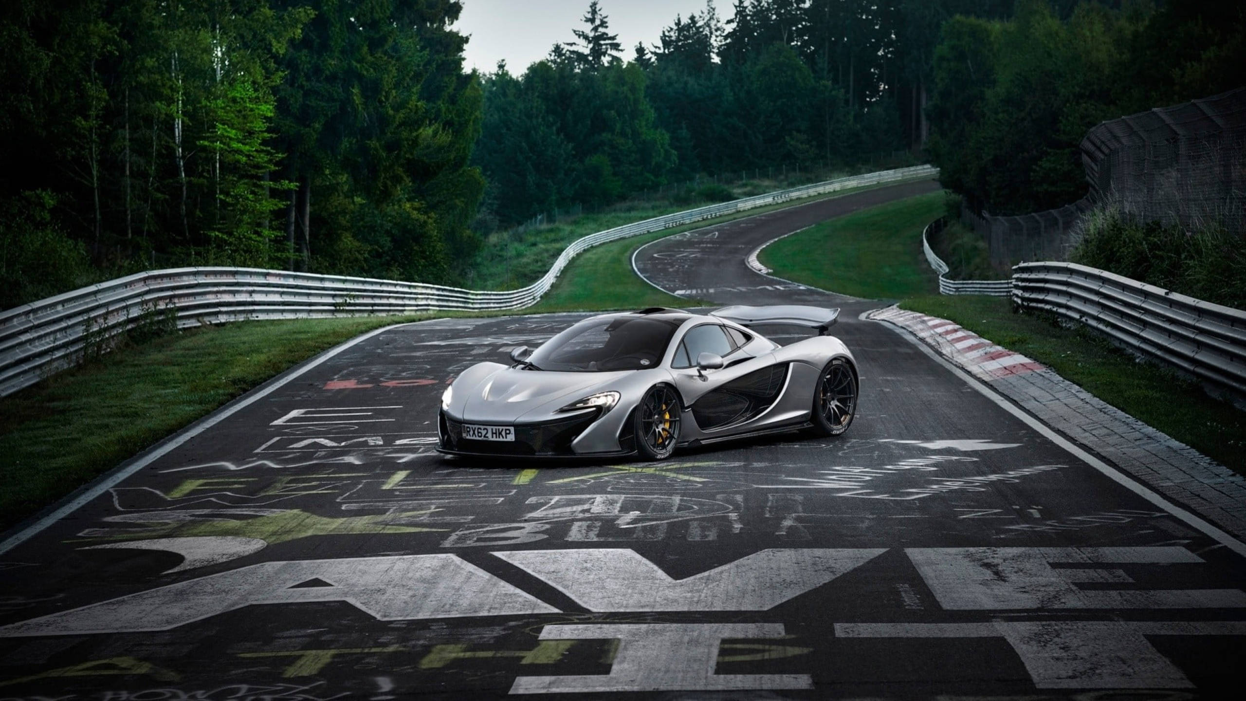 賽車 桌布 壁紙 — 一架流線型的銀色McLaren P1跑車在蜿蜒的瀝青賽道上行駛，周圍是茂密的綠色森林。