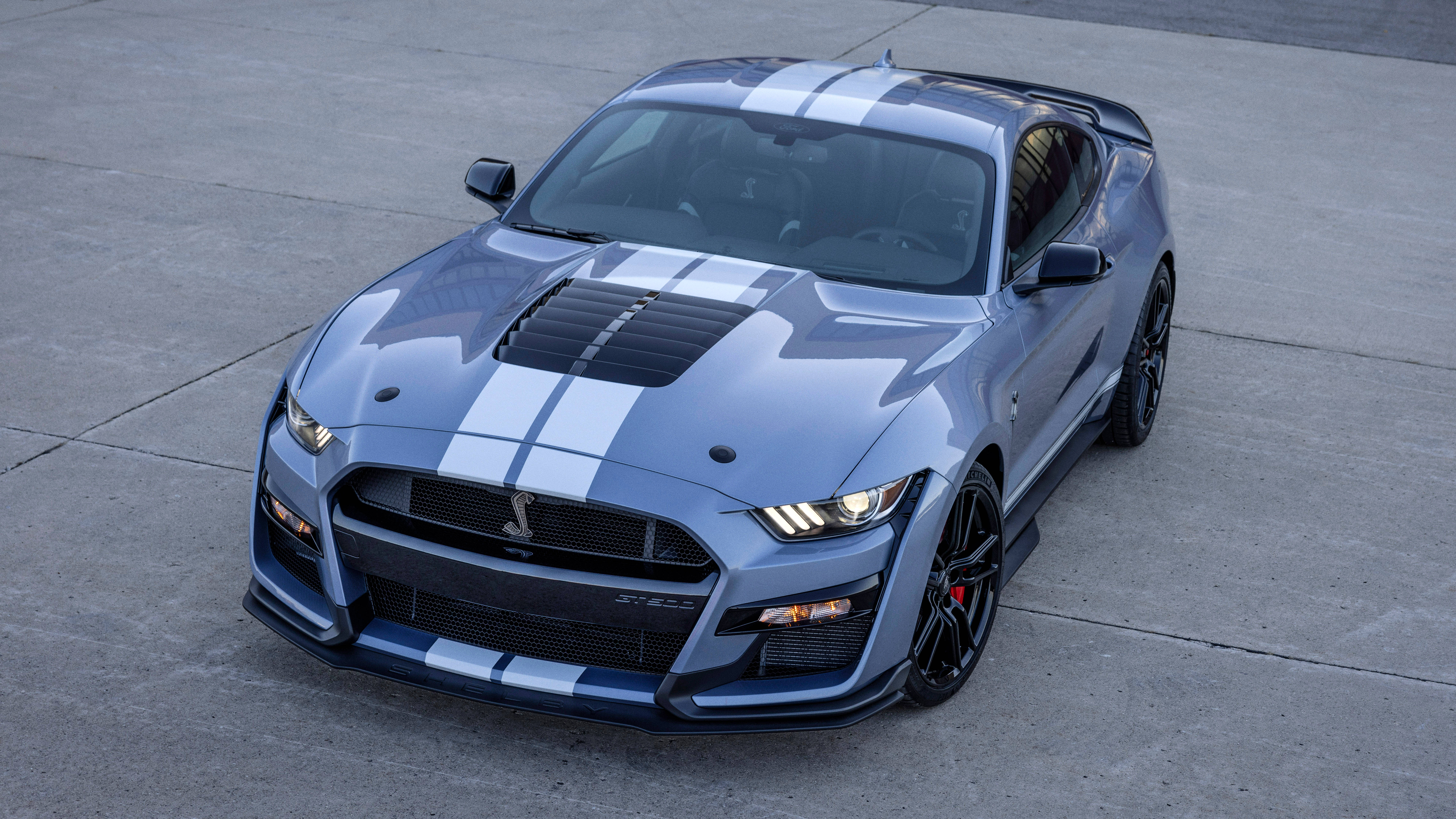 野馬GIF 桌布 壁紙 — 一架流線型的銀色Shelby GT500，帶有醒目的白色賽車條紋和黑色裝飾，在明亮的日光下在混凝土表面上閃閃發光。