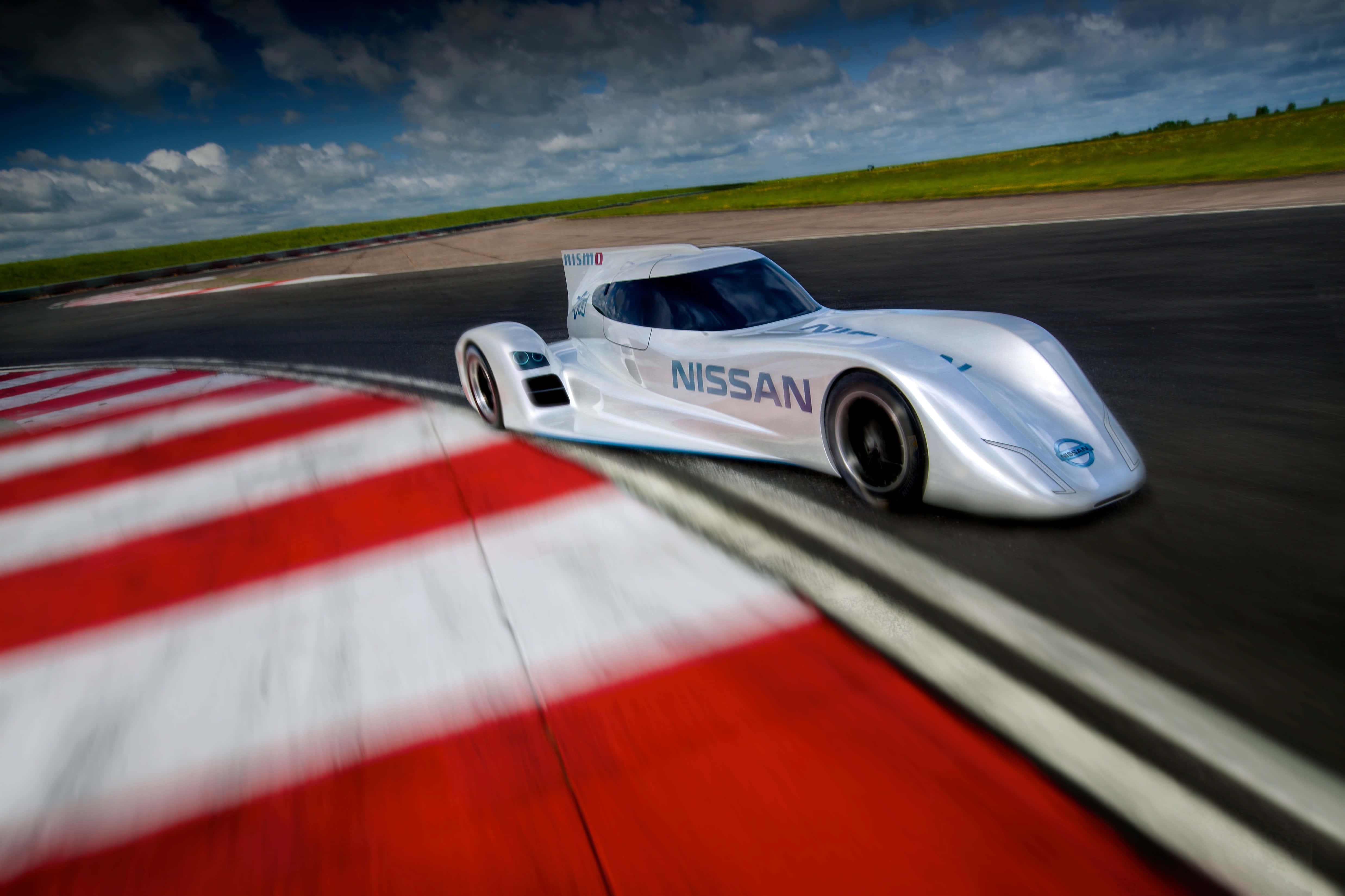Car Racing 桌布 壁紙 — 一架流線型的銀色日產賽車在陰天之下，於設有紅白路緣石的賽道彎道上疾馳。