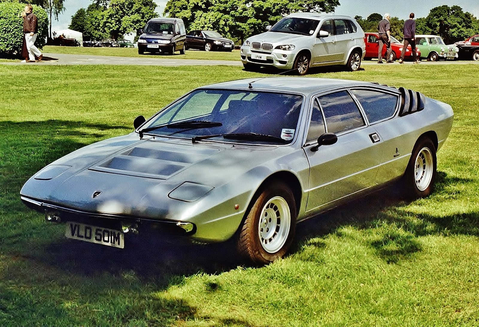 Lamborghini Urraco 桌布 壁紙 — 一架流線型的銀色藍寶堅尼Countach停放在經典車展的翠綠草地上，背景有其他車輛和人群。