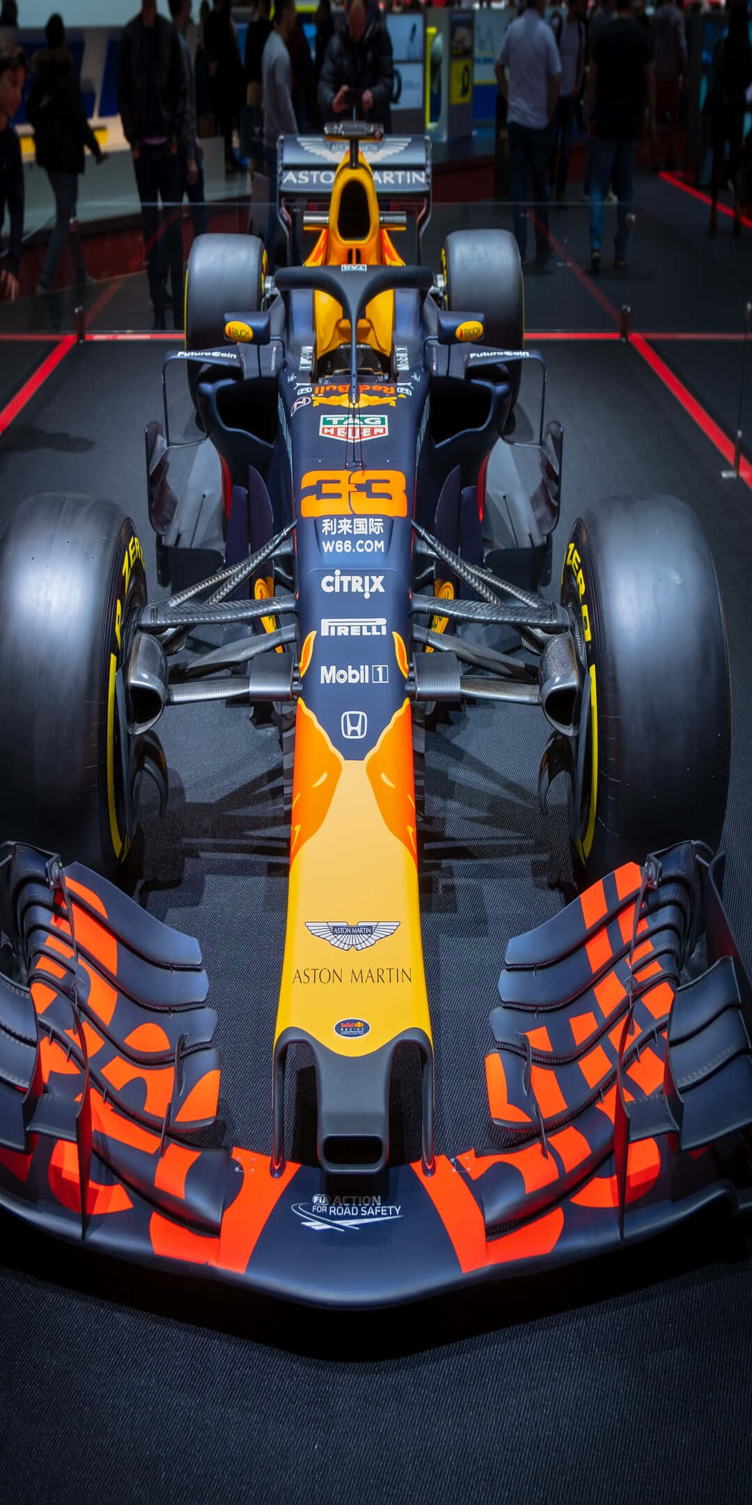 F1 2016 桌布 壁紙 — 一架流線型的阿斯頓馬丁一級方程式賽車，帶有鮮豔的橙色和黑色塗裝，在車展的深色地板上展示。