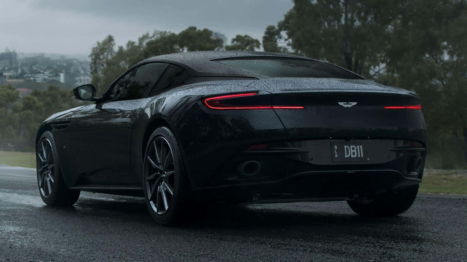 Aston Martin Db11 桌布 壁紙 — 一架流線型的黑色Aston Martin DB11在車尾閃爍著雨滴，紅色尾燈在霧濛濛的郊區背景下發光。