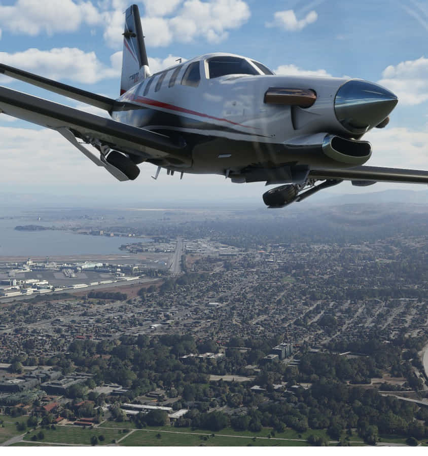 Android Microsoft Flight Simulator 桌布 壁紙 — 一架流線型私人飛機在晴朗的藍天下，翱翔於擁有海岸灣和遠山的廣闊城市景觀之上。