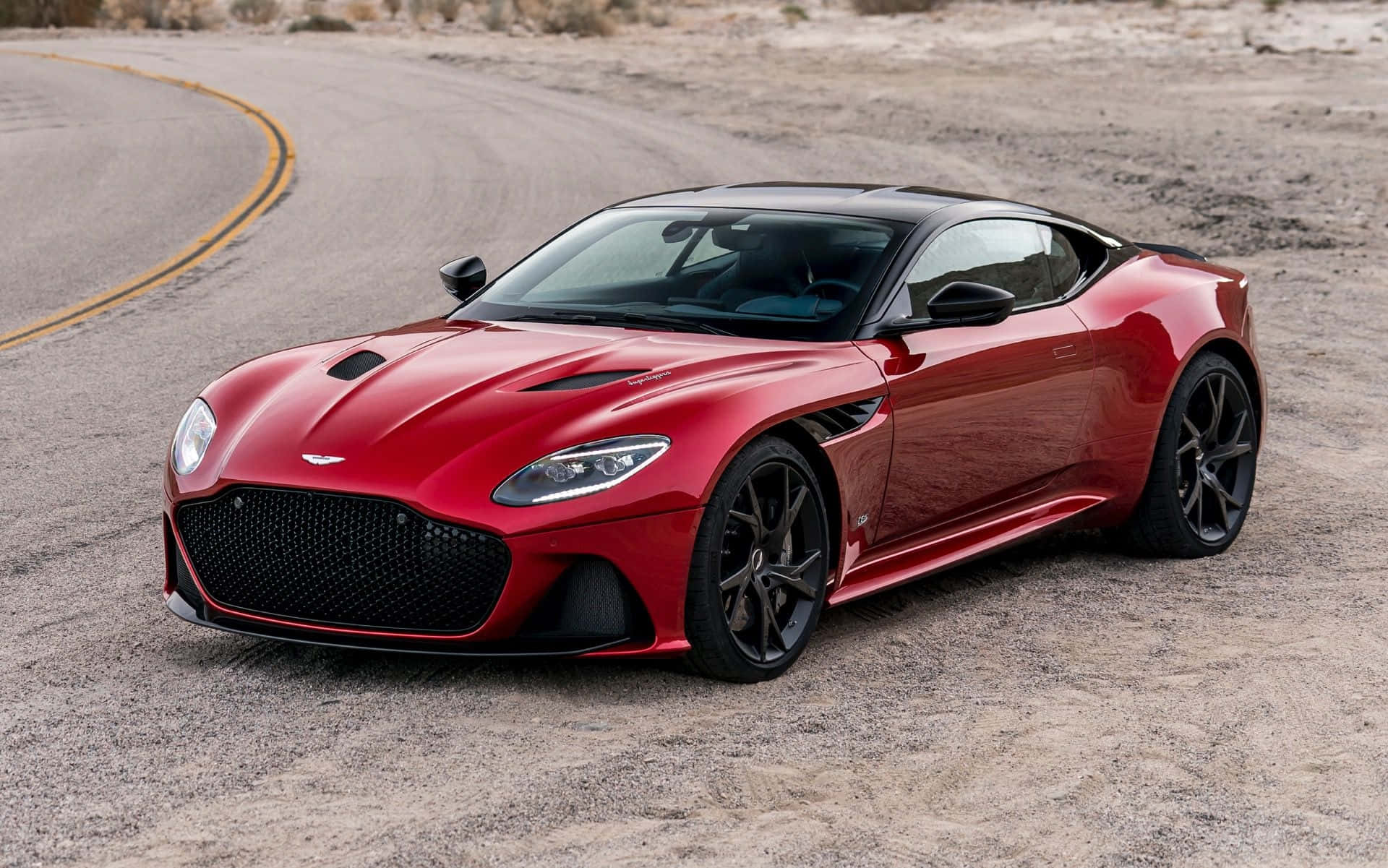 奧斯頓馬丁 桌布 壁紙 — 一架流線型紅色Aston Martin DBS Superleggera停放在蜿蜒的沙漠公路上，背景是崎嶇的地形。