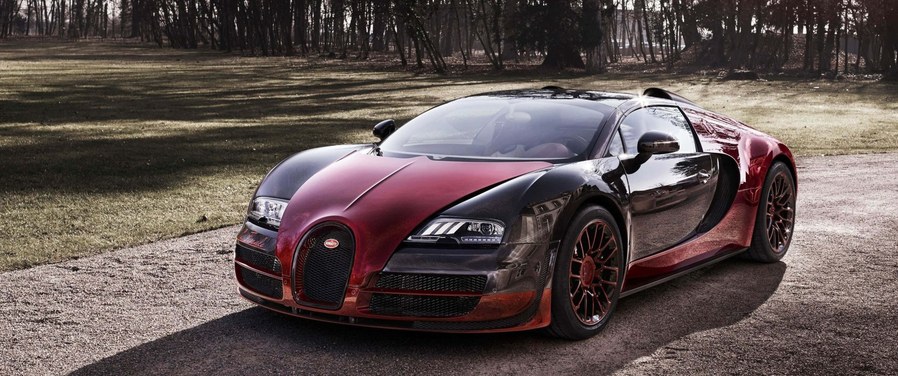3440x1440p Bugatti 桌布 壁紙 — 一架流線型紅黑色布加迪威龍停泊在碎石路上，旁邊是草地，背景有樹木。