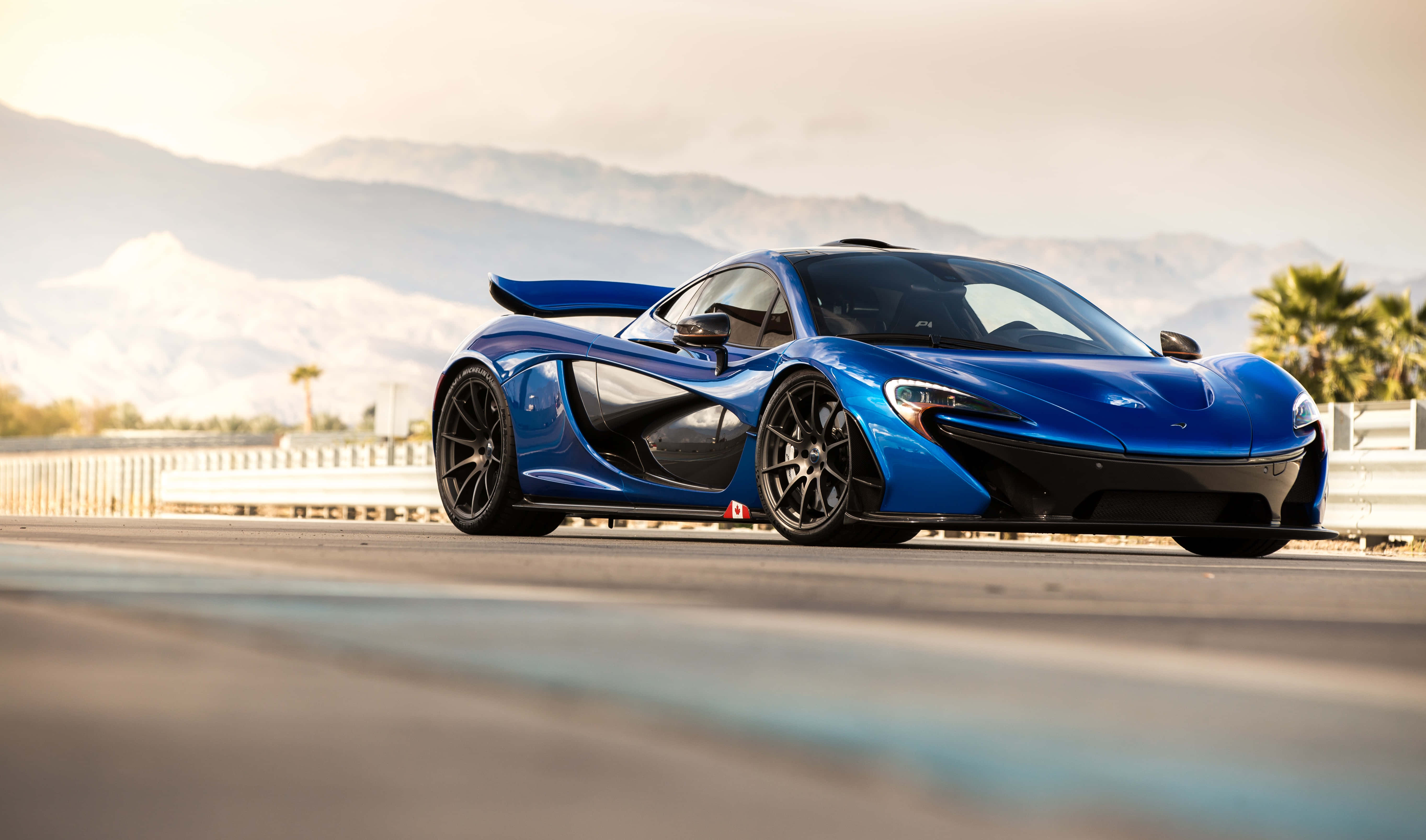 麥拉倫 桌布 壁紙 — 一架流線型藍色McLaren P1跑車在沙漠公路上滑行，遠方有山脈和棕櫚樹，天空朦朧。