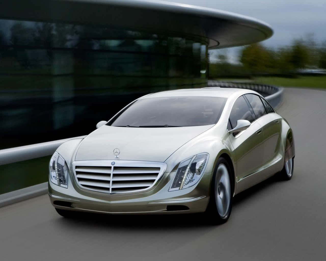 跑車 桌布 壁紙 — 一架流線型銀色 Mercedes-Benz 概念車在陰天下一條彎曲道路上行駛，旁邊是一座現代玻璃建築。