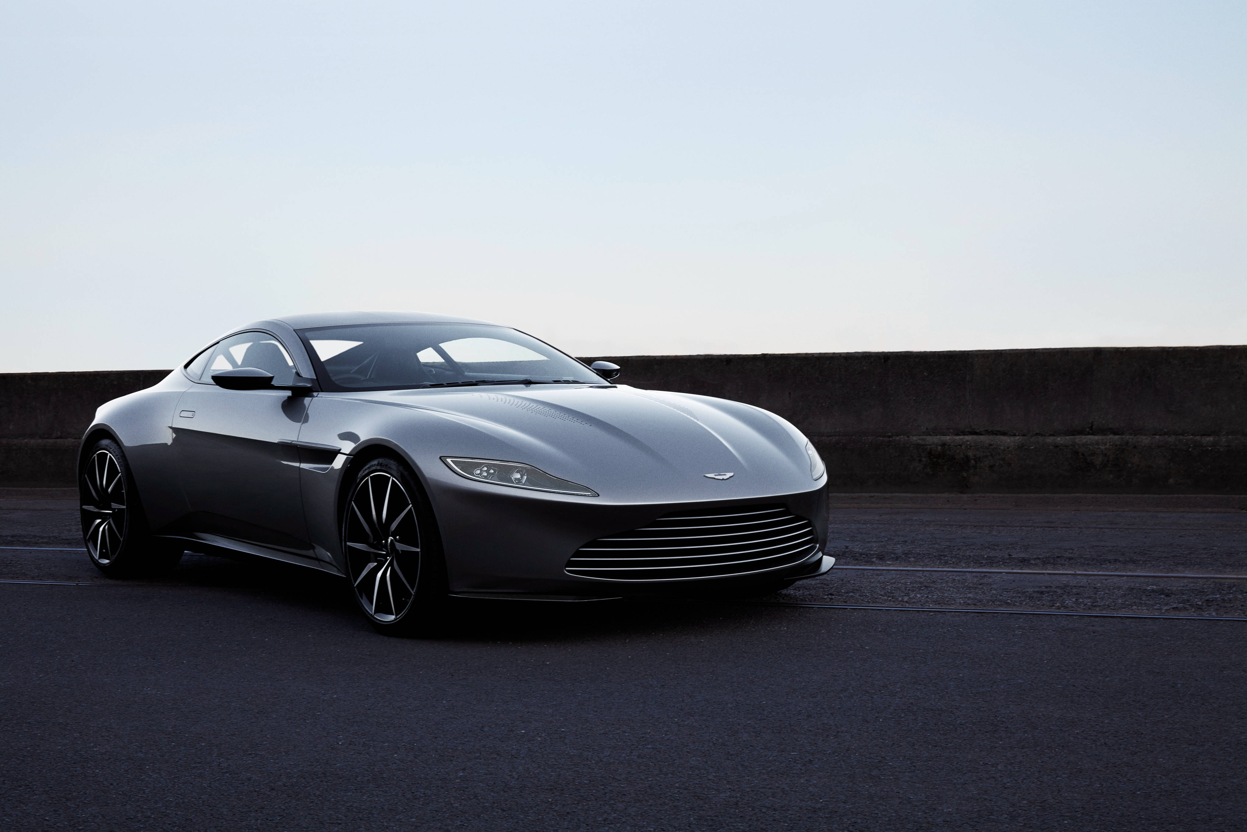銀色 桌布 壁紙 — 一架流線型銀色Aston Martin跑車停泊在灰濛天空下的深色瀝青路上，旁邊是混凝土護欄。