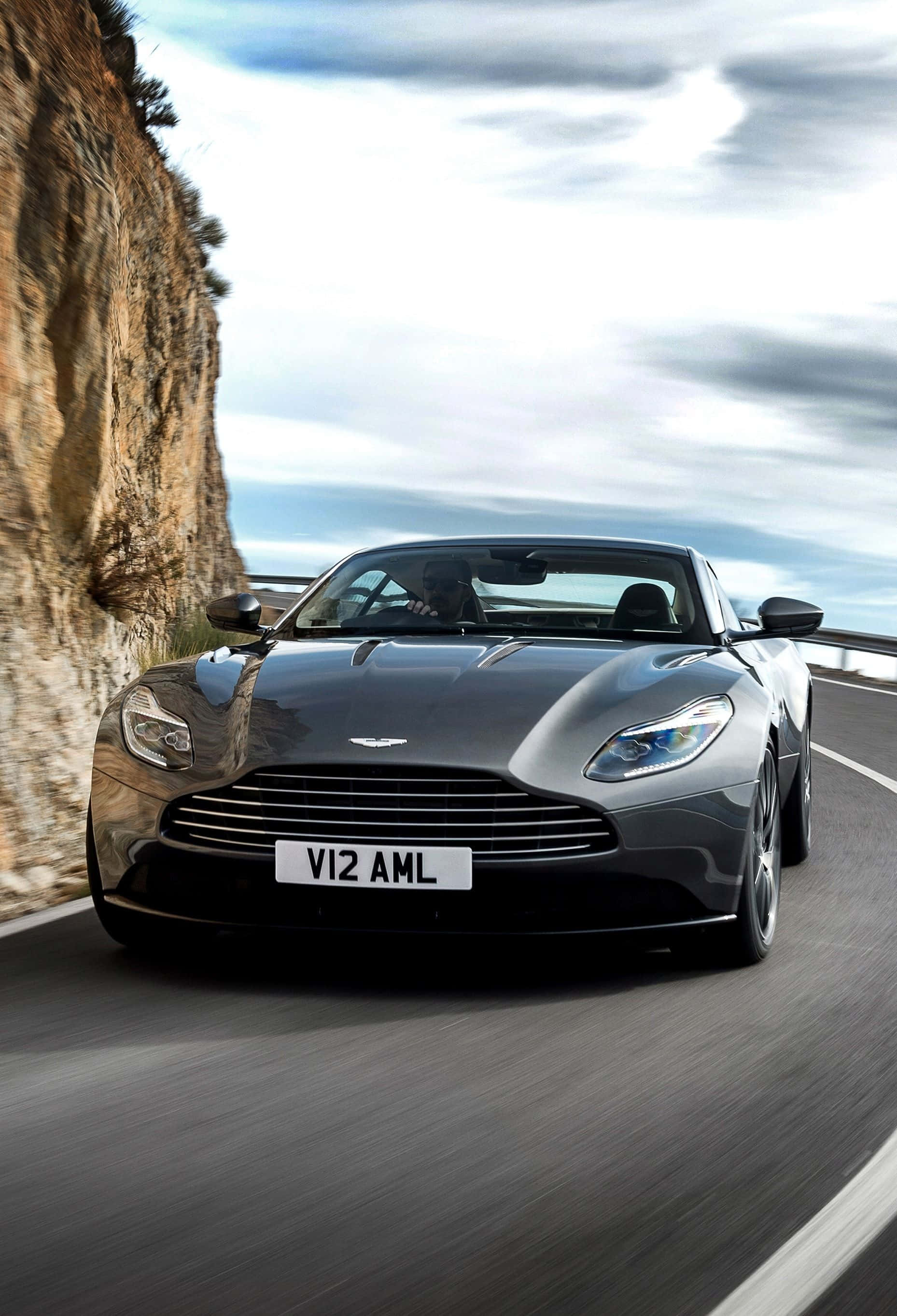 Aston Martin Rapide S 桌布 壁紙 — 一架流線型銀色Aston Martin V12敞篷跑車，在多雲的天空下，沿著崎嶇懸崖旁蜿蜒的海岸公路奔馳。