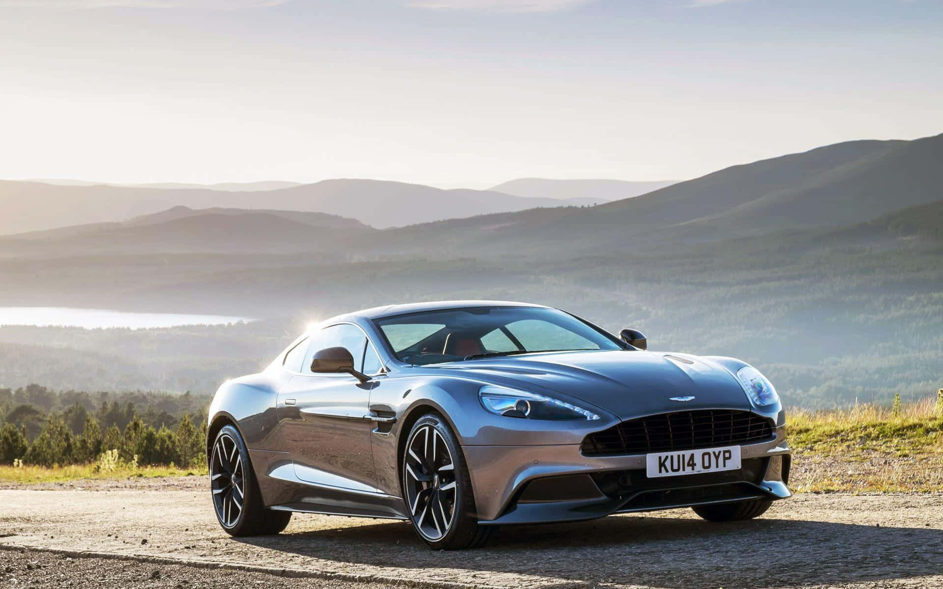 奧斯頓馬丁 桌布 壁紙 — 一架流線型銀色Aston Martin跑車停泊在碎石路上，背景是起伏的山丘和遠處的湖泊，天空柔和晨光。