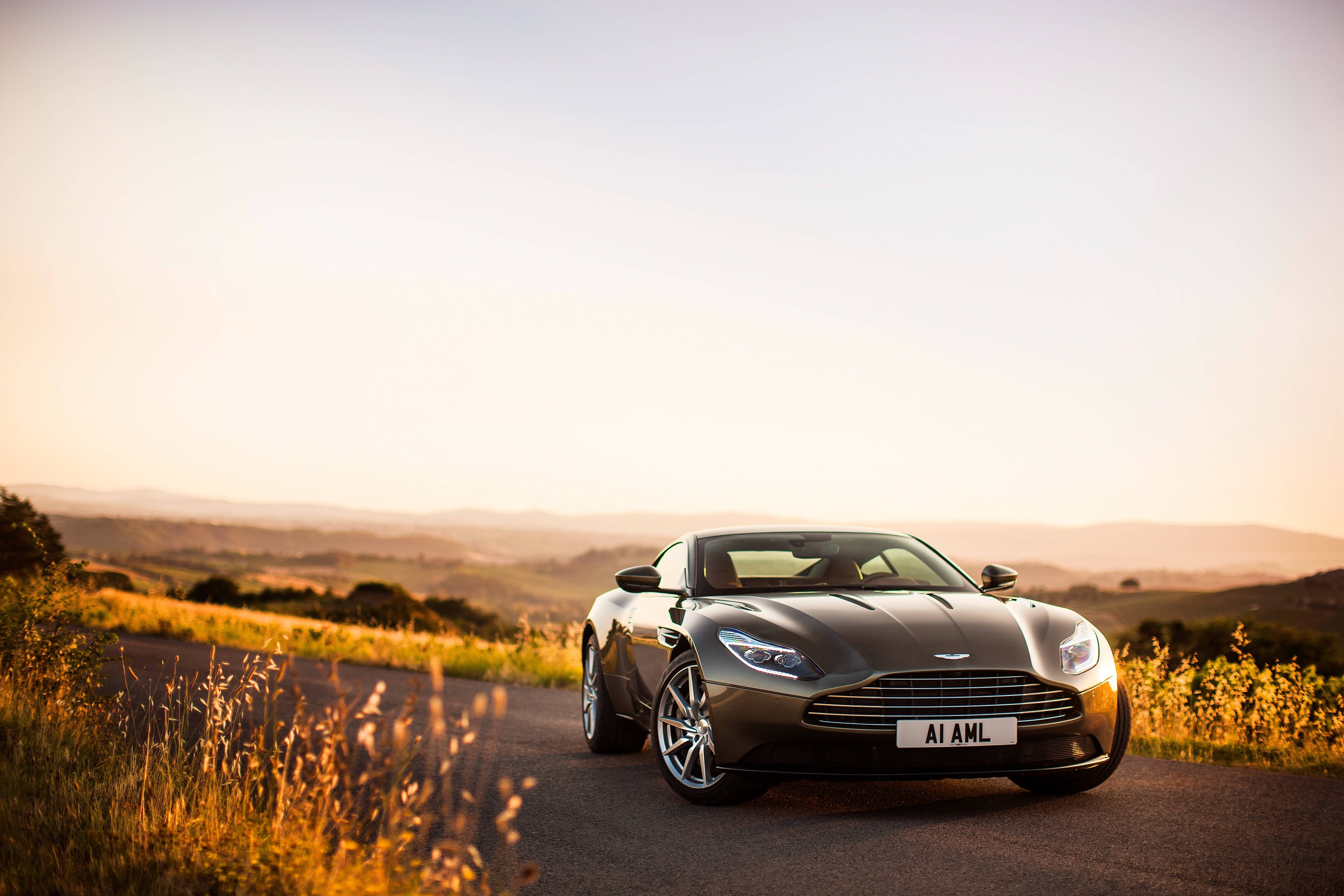 奧斯頓馬丁 桌布 壁紙 — 一架流線型銀色Aston Martin DB11停泊在蜿蜒的鄉村道路上，背景是金色夕陽和起伏的山丘。