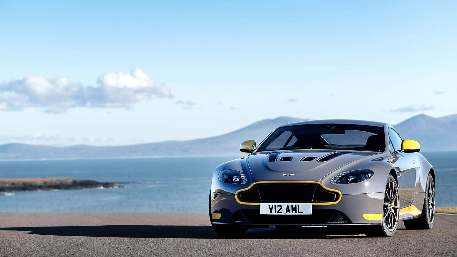 Aston Martin V12 Vantage 桌布 壁紙 — 一架流線型銀色Aston Martin跑車，帶有黃色點綴，停泊在沿海公路上，俯瞰著海洋和遠處的山脈，天空湛藍明亮。